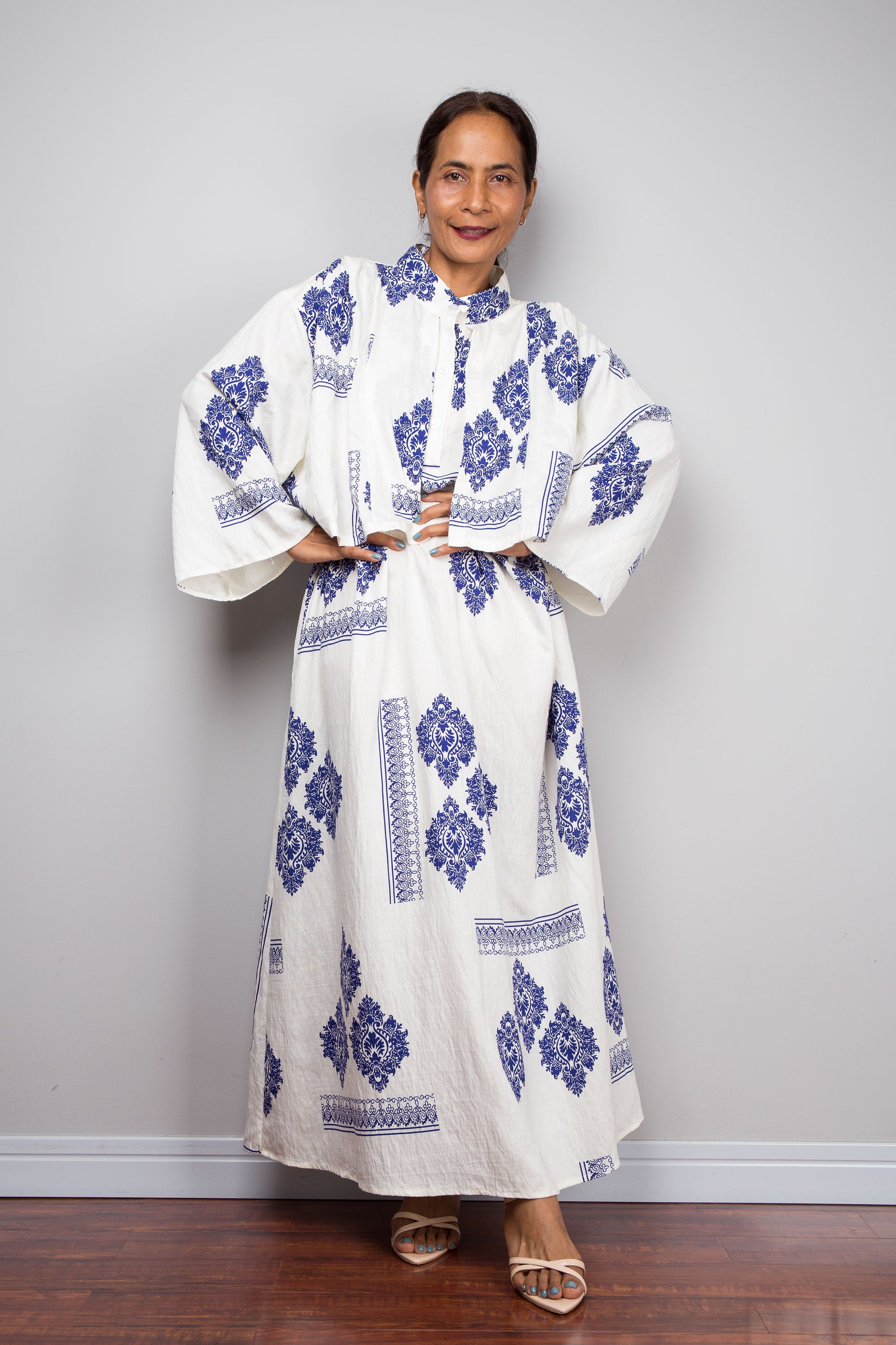 A woman modeling a white caftan maxi dress with blue floral and geometric patterns, standing against a light gray wall on a wooden floor.