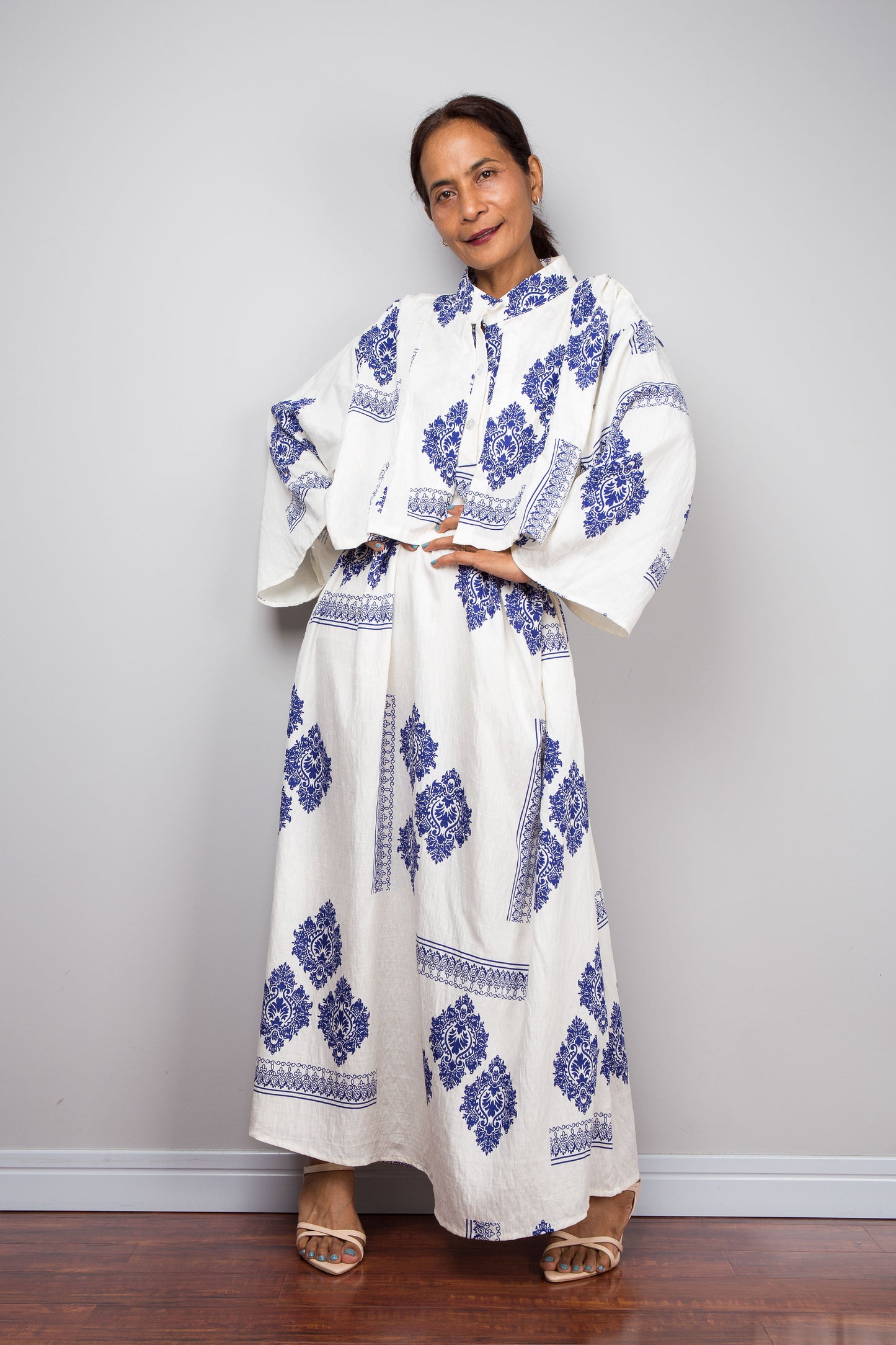 A woman modeling a long off white maxi caftan dress with blue floral and geometric patterns, standing against a light gray wall on a dark wood floor.