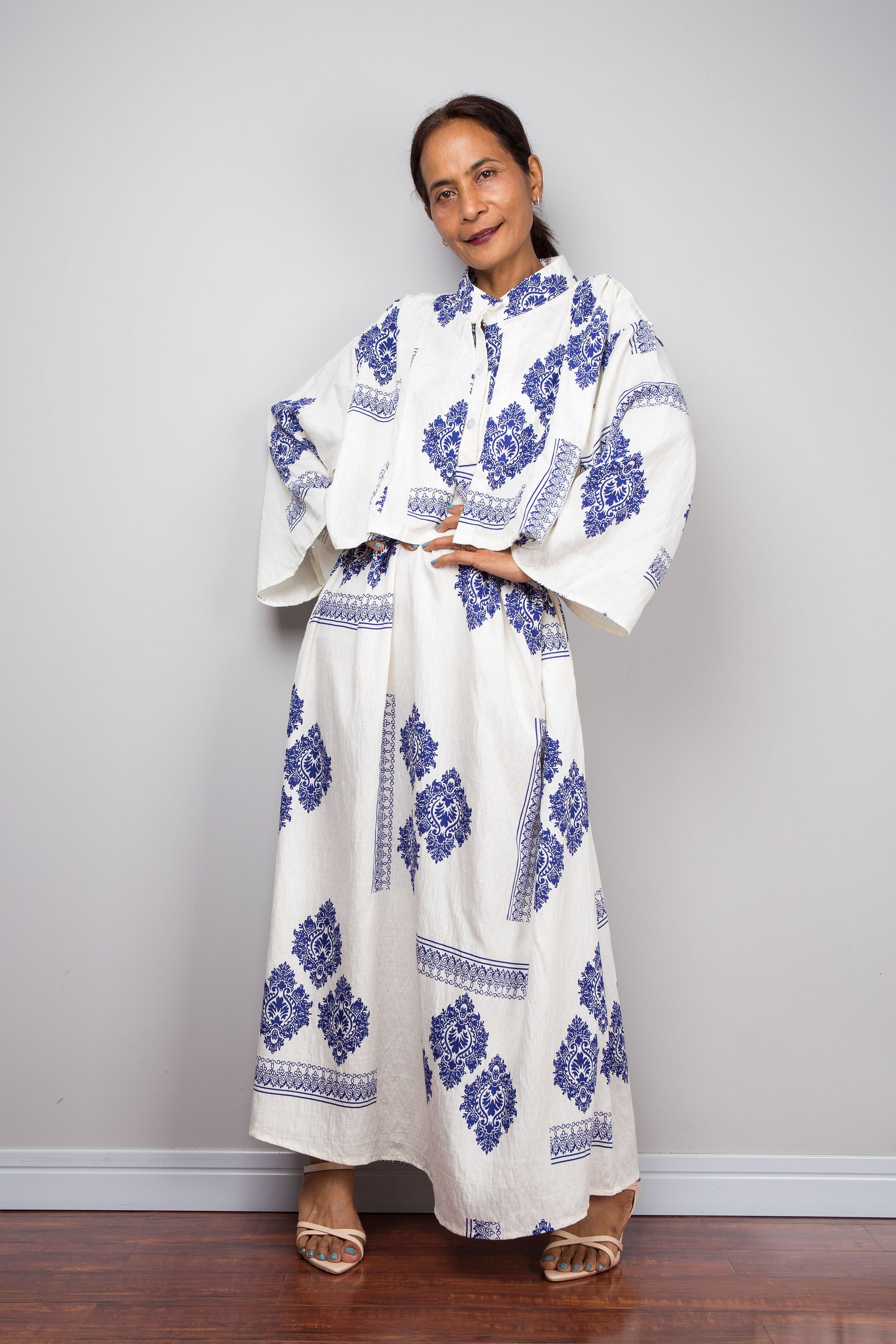 A woman modeling a long off white maxi caftan dress with blue floral and geometric patterns, standing against a light gray wall on a dark wood floor.