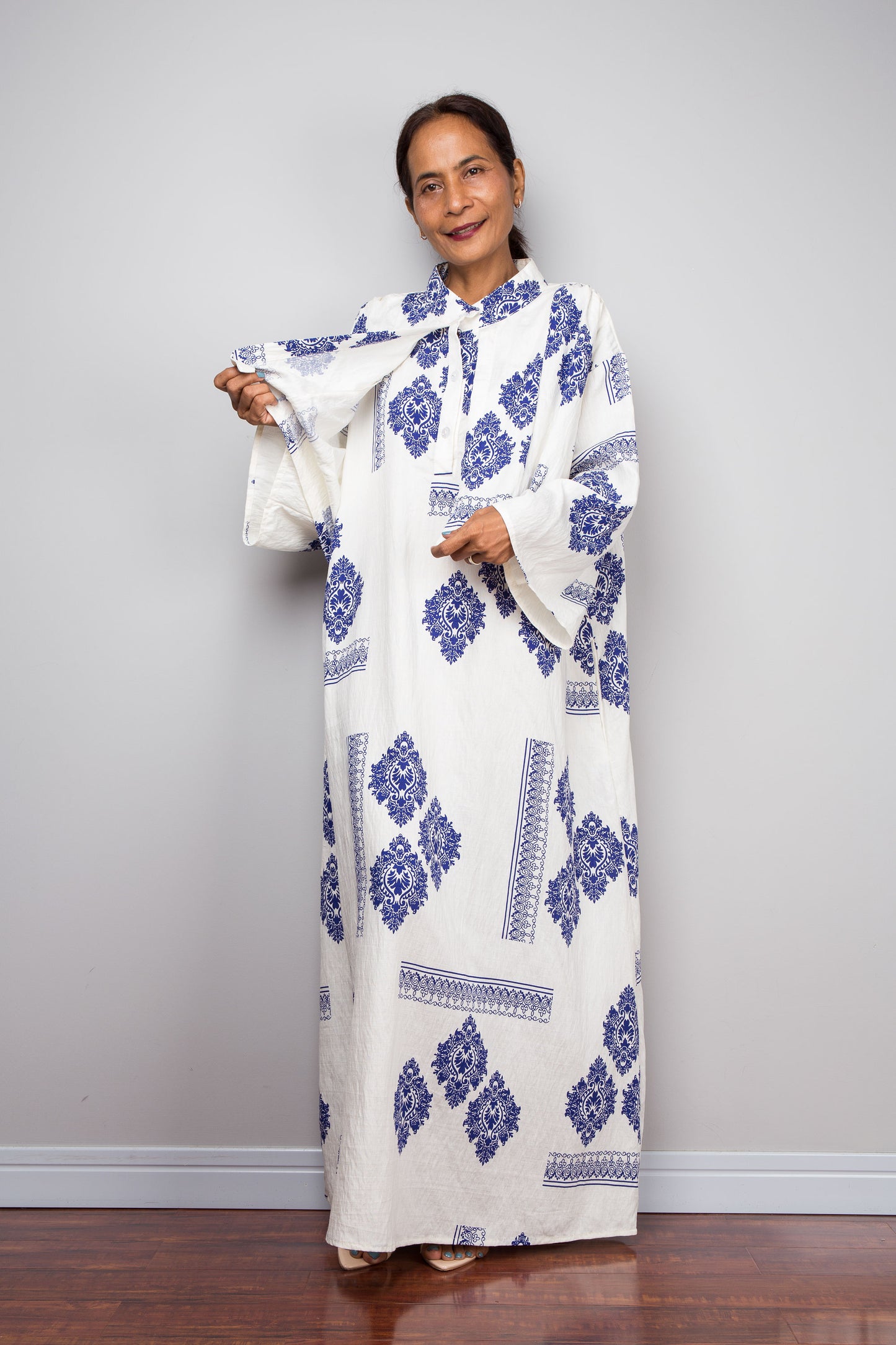 Front view of a female model wearing the Nuichan white and blue medallion kaftan dress while lifting the attached capelet to demonstrate its construction as a separate freely draping layer, displaying the full body of the dress beneath with diamond medallion print and decorative border banding