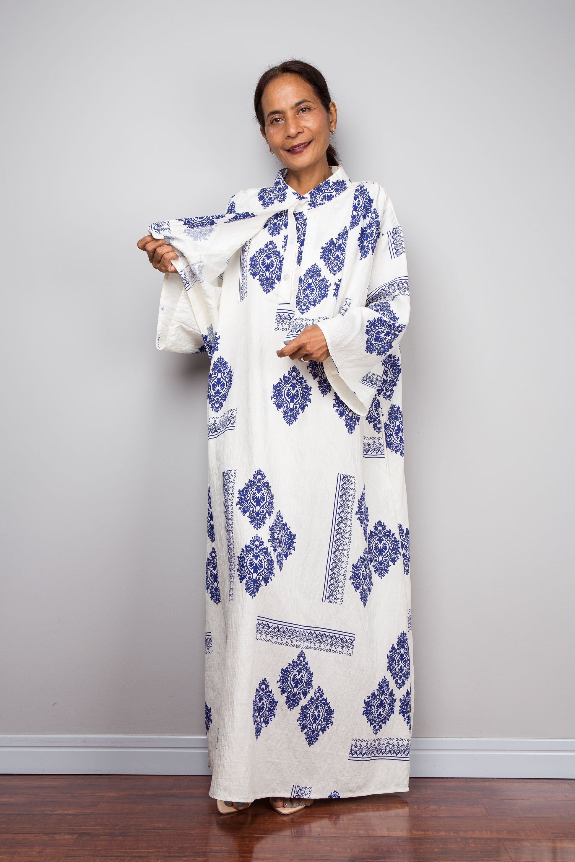 Front view of a female model wearing the Nuichan white and blue medallion kaftan dress while lifting the attached capelet to demonstrate its construction as a separate freely draping layer, displaying the full body of the dress beneath with diamond medallion print and decorative border banding