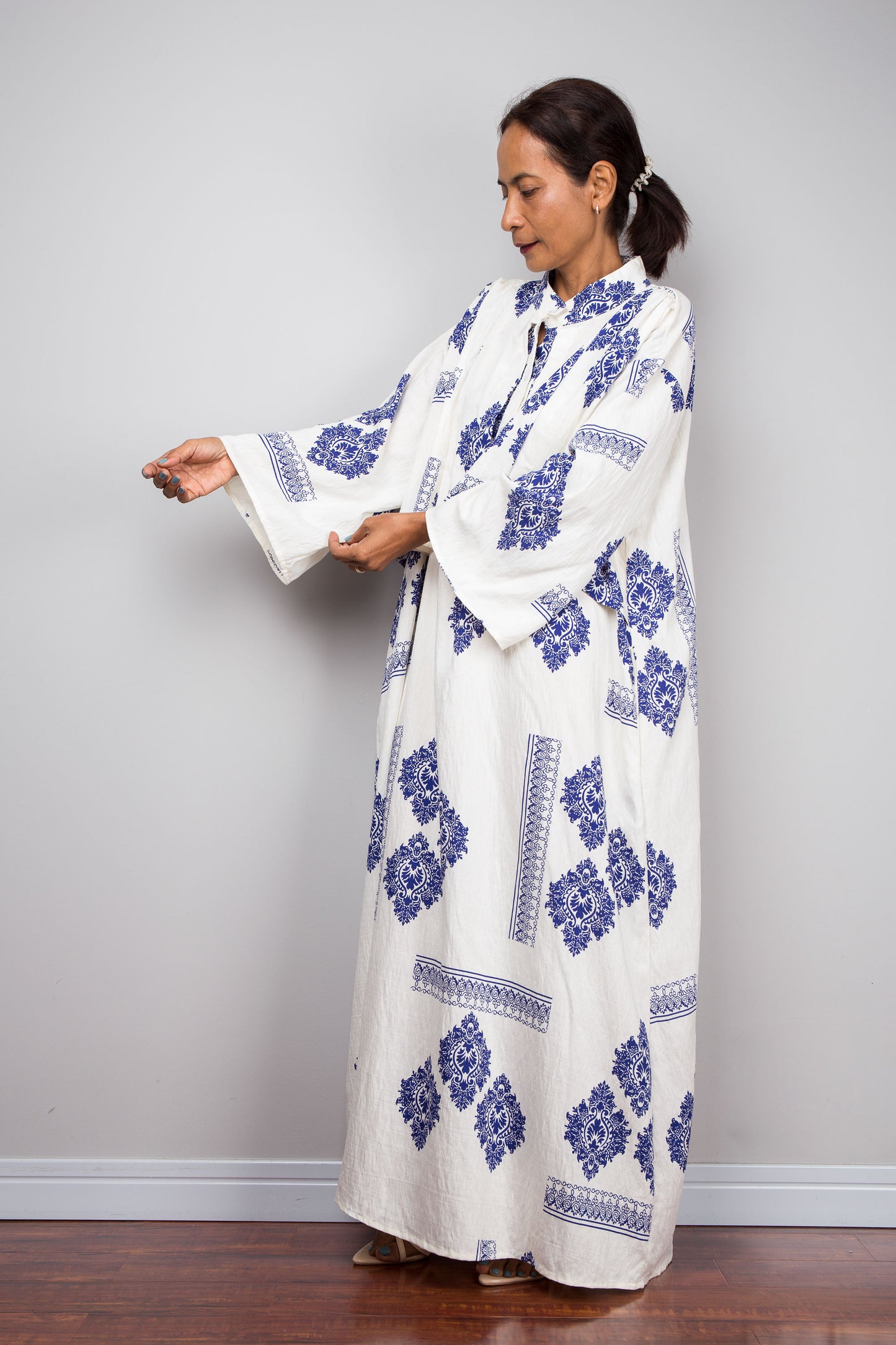 A woman wearing a long white caftan dress with blue floral and geometric block prints, standing in a studio setting with a gray wall and wooden floor.