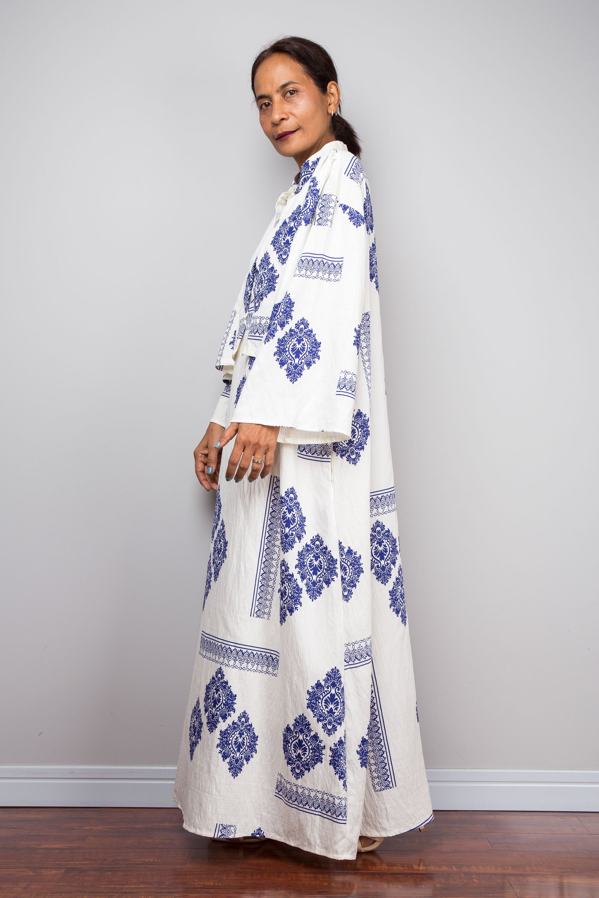 Side view of A woman wearing a long white kaftan dress with blue geometric and floral patterns, standing in profile against a light gray wall.