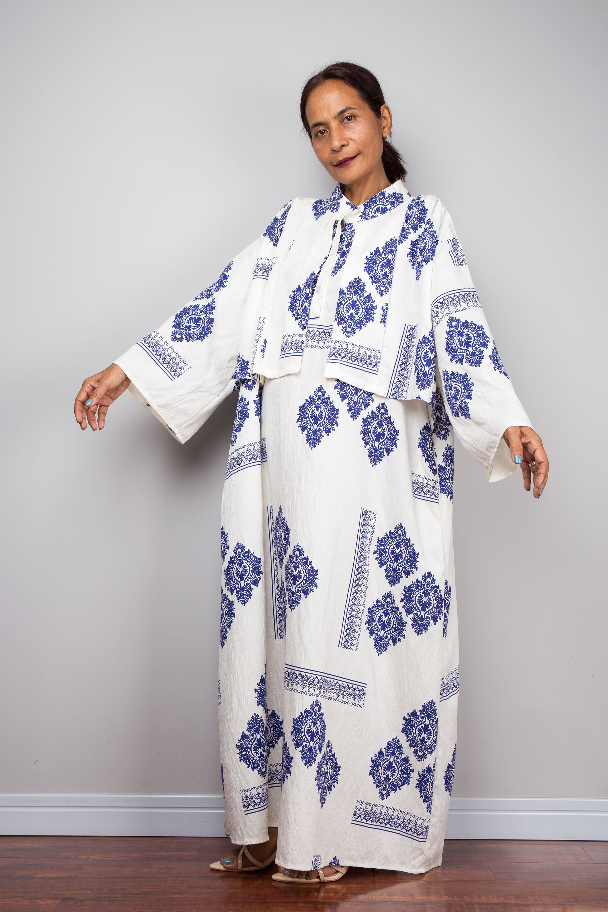 Three-quarter side view of a model wearing the Nuichan white and blue medallion print capelet kaftan maxi dress with arms relaxed, displaying the full floor-length A-line silhouette, freely draping attached capelet, mandarin collar, flared sleeve cuffs with blue border detail, and cobalt blue medallion placement print throughout
