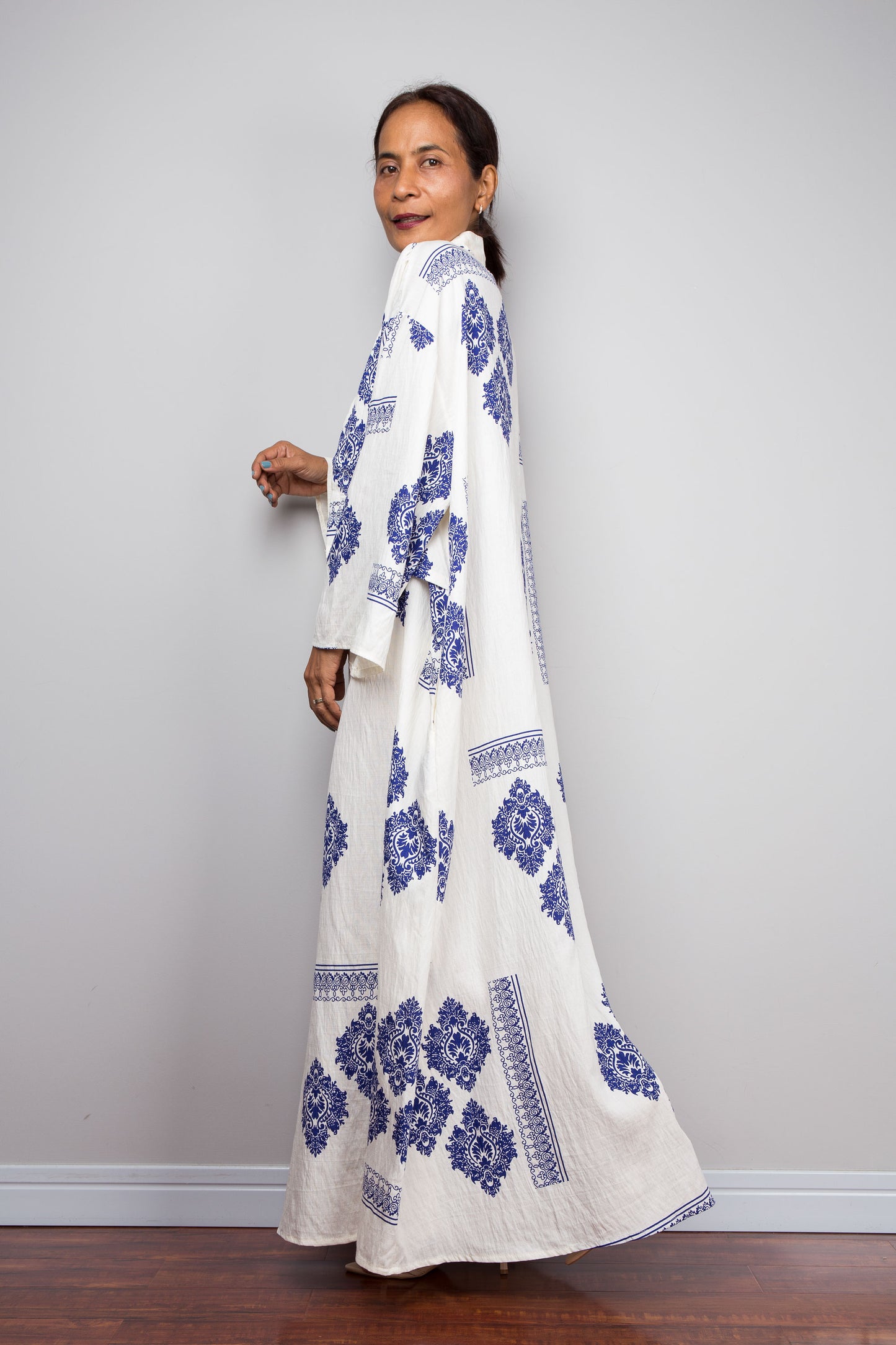 A woman modeling a long white maxi caftan dress with blue floral block prints, standing against a light gray wall on a dark wood floor.