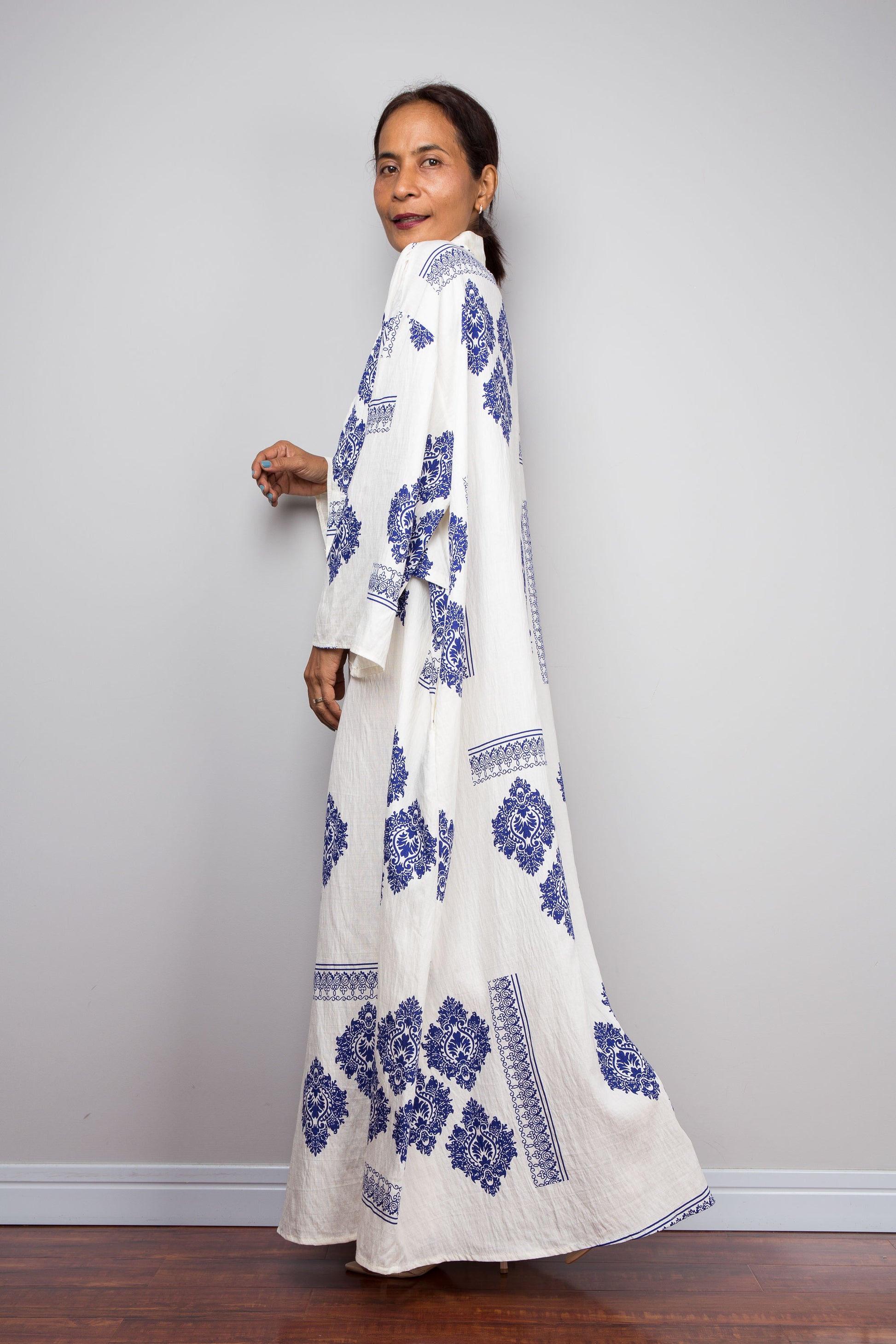A woman modeling a long white maxi caftan dress with blue floral block prints, standing against a light gray wall on a dark wood floor.