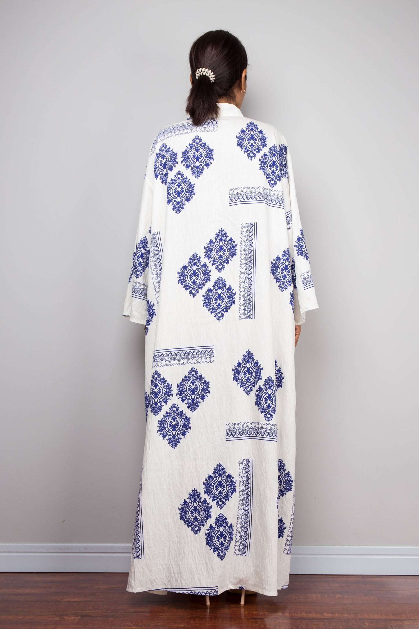A woman viewed from behind wearing a long white kaftan-style dress with blue geometric and floral embroidery, standing against a plain light gray wall.