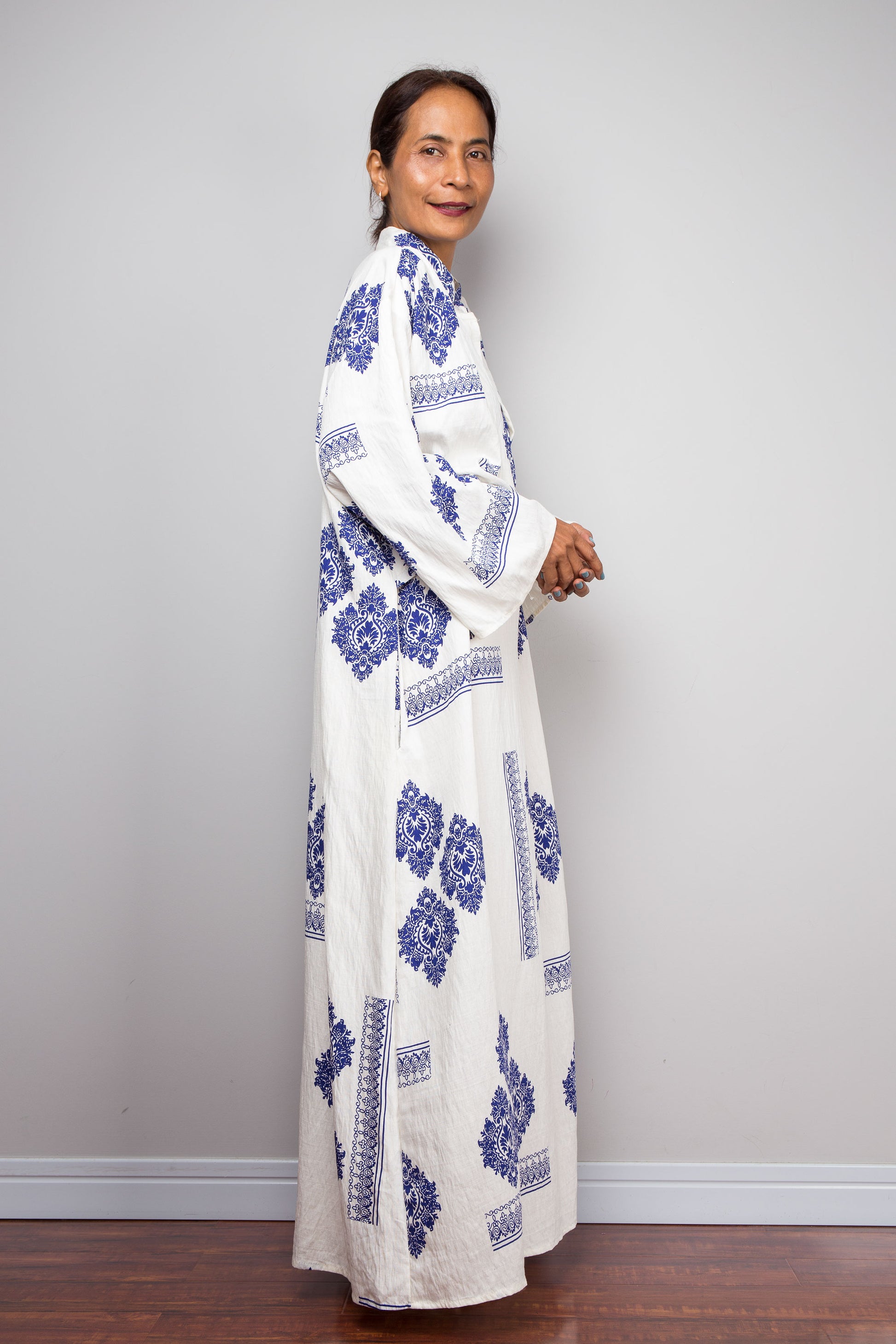 A woman modeling a long white kaftan dress with blue floral and geometric block-print patterns, standing against a light gray wall.