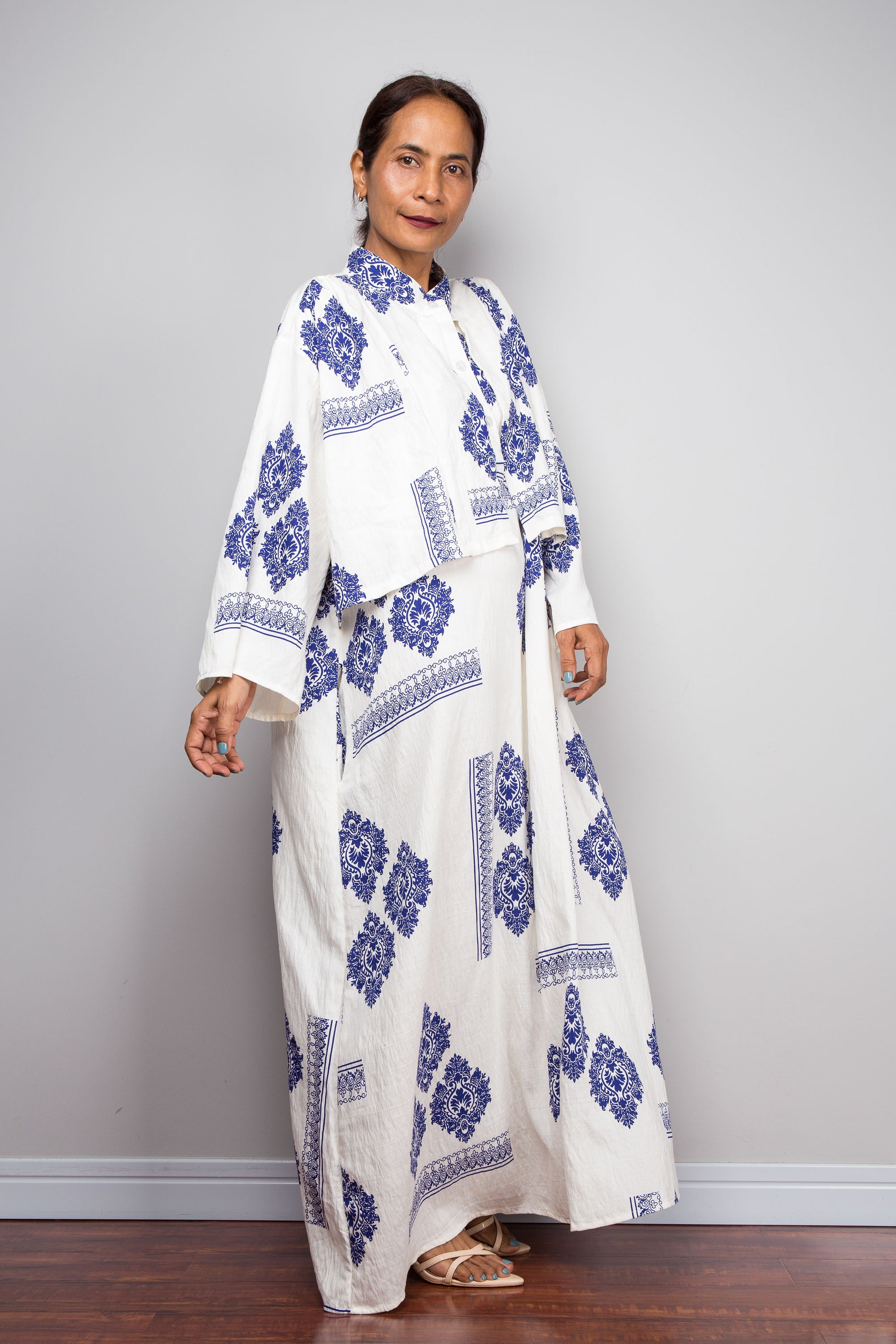 A woman modeling a white caftan dress with blue floral and geometric block prints, against a plain gray wall.