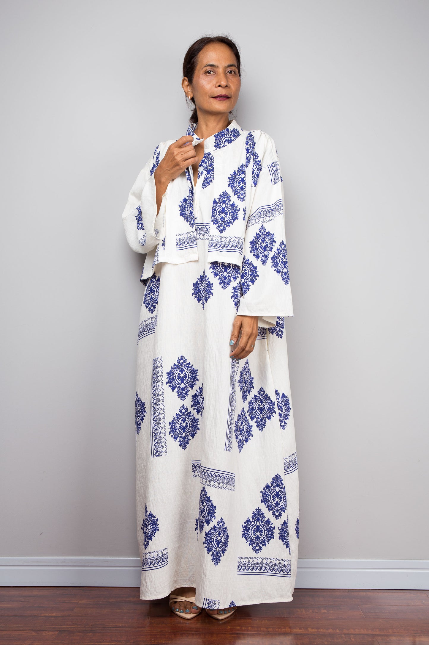 A woman modeling a white traditional long-sleeve caftan with blue geometric block prints, standing against a light gray wall and dark wood floor.