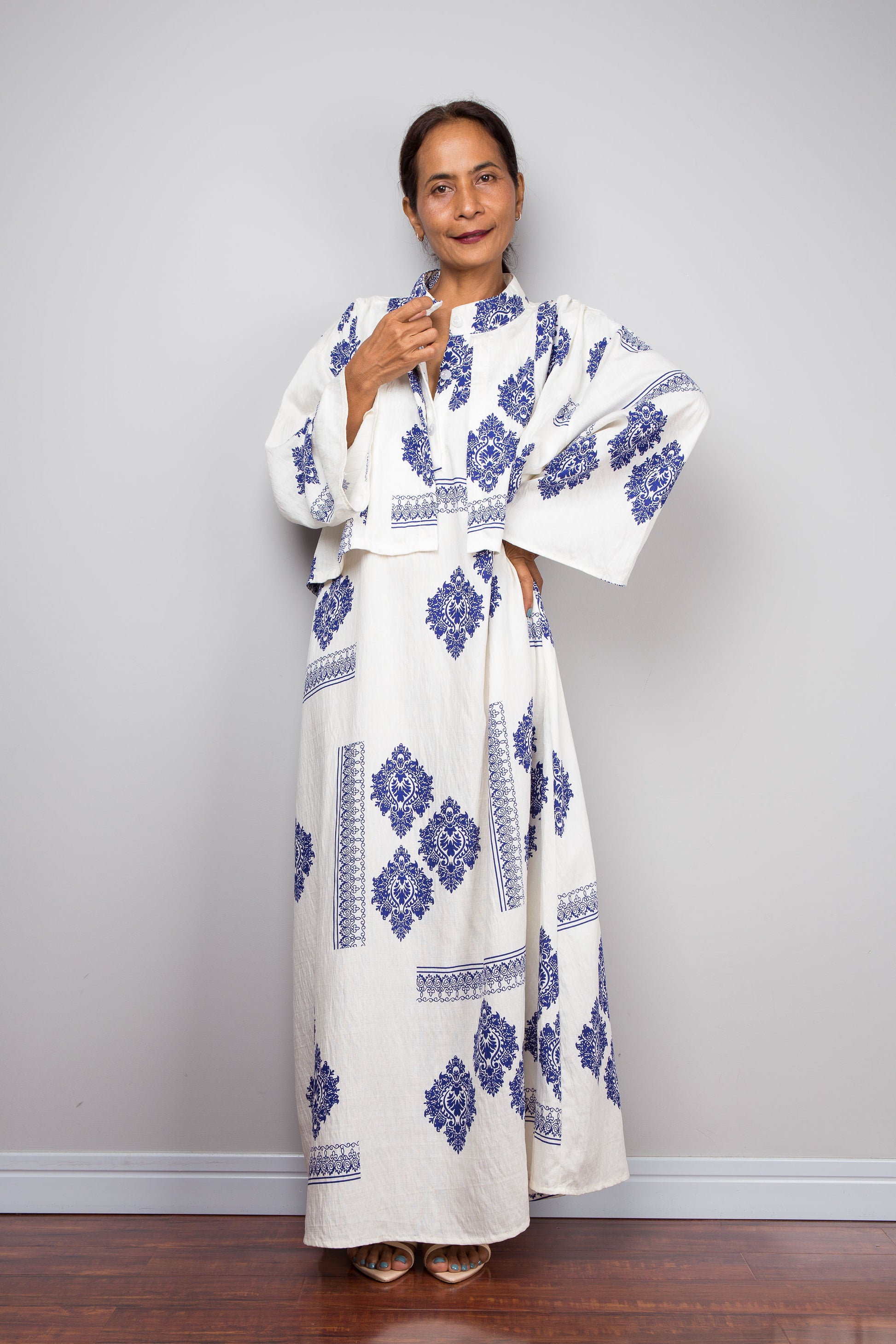 Front view of a model wearing the Nuichan white and blue medallion kaftan dress while lifting the attached capelet to demonstrate its construction as a separate freely draping layer, displaying the full body of the dress beneath with diamond medallion print and decorative border banding