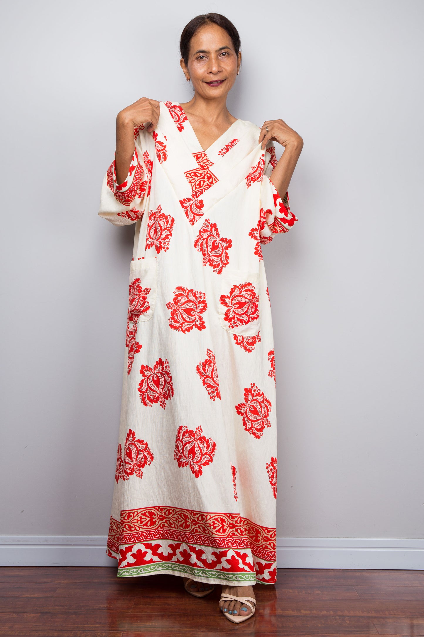 Woman wearing a long off white bohemian caftan dress with red floral patterns against a plain background