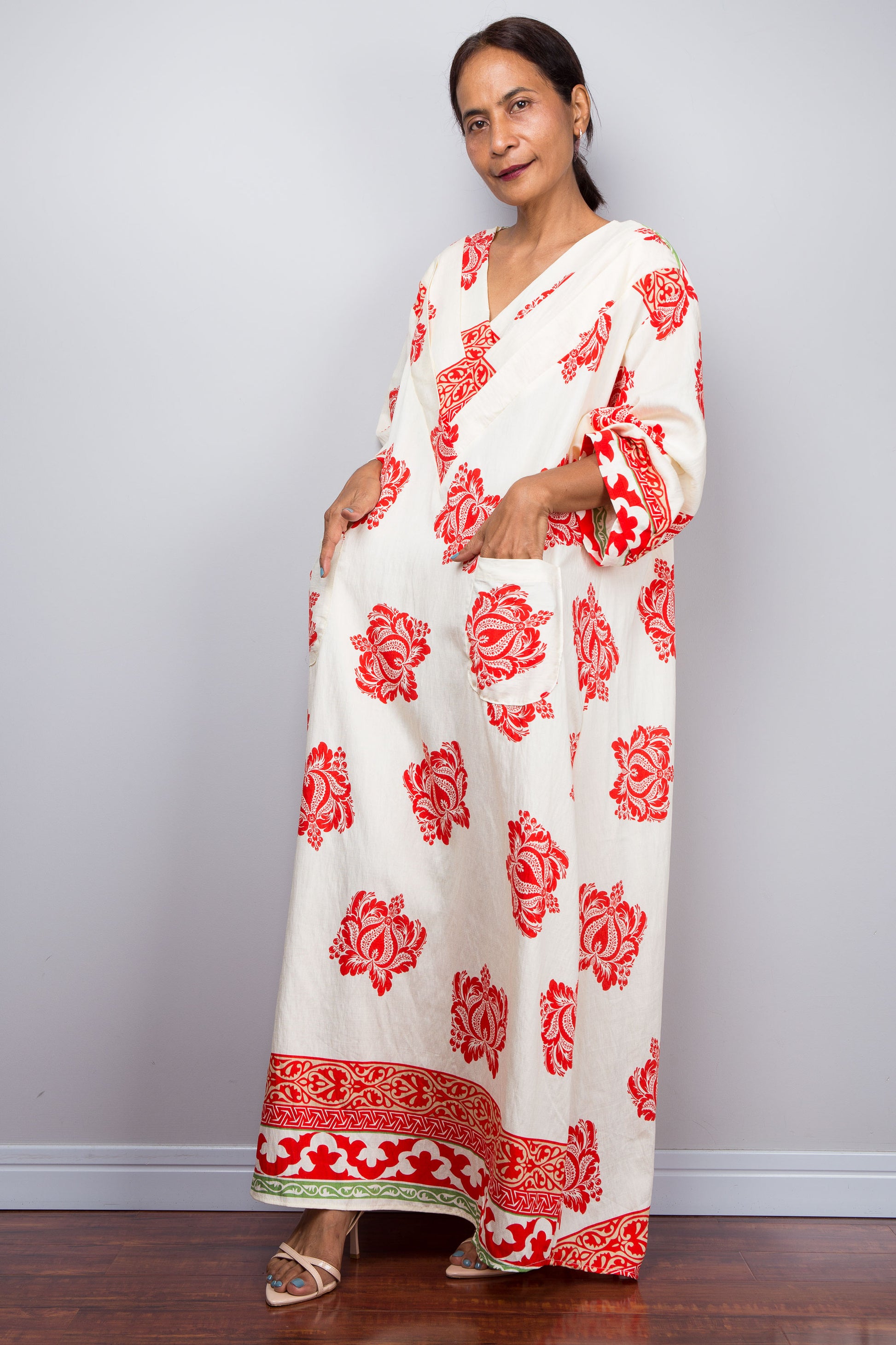 Woman wearing an off white kaftan dress with red floral patterns against a plain background