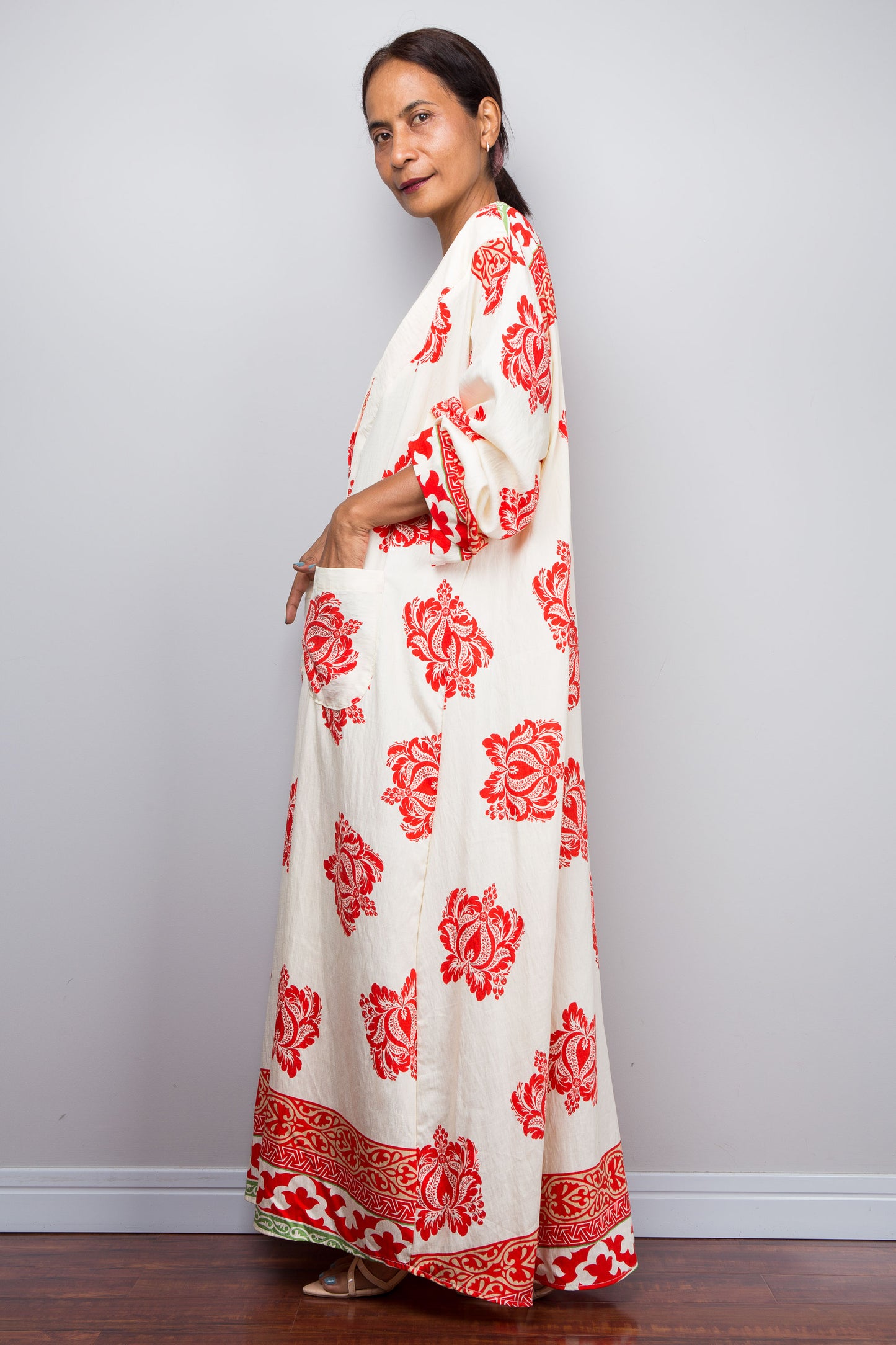 Woman wearing a long, white dress with red floral patterns, modeling the pockets, against a plain background