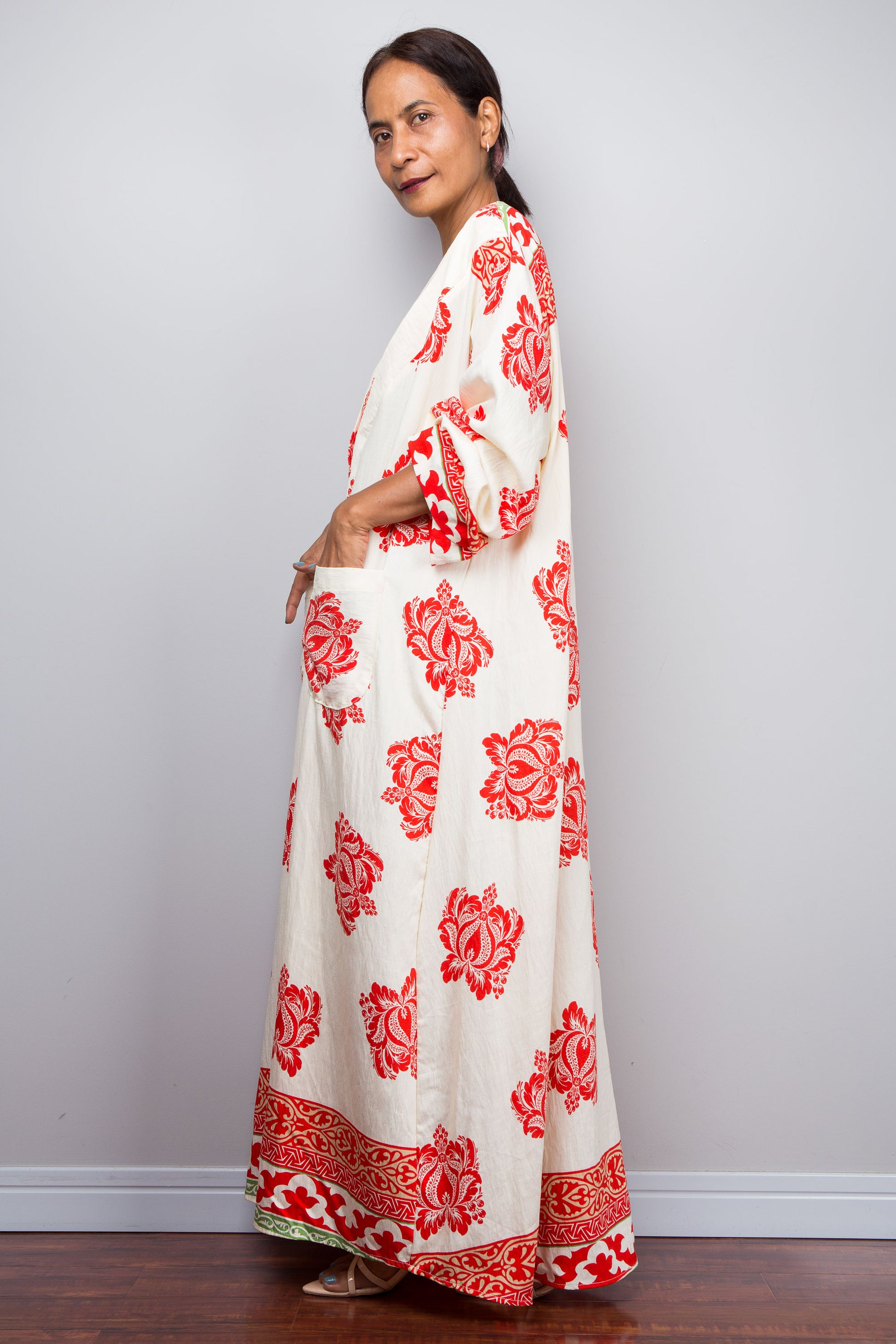 Woman wearing a long, white dress with red floral patterns, modeling the pockets, against a plain background