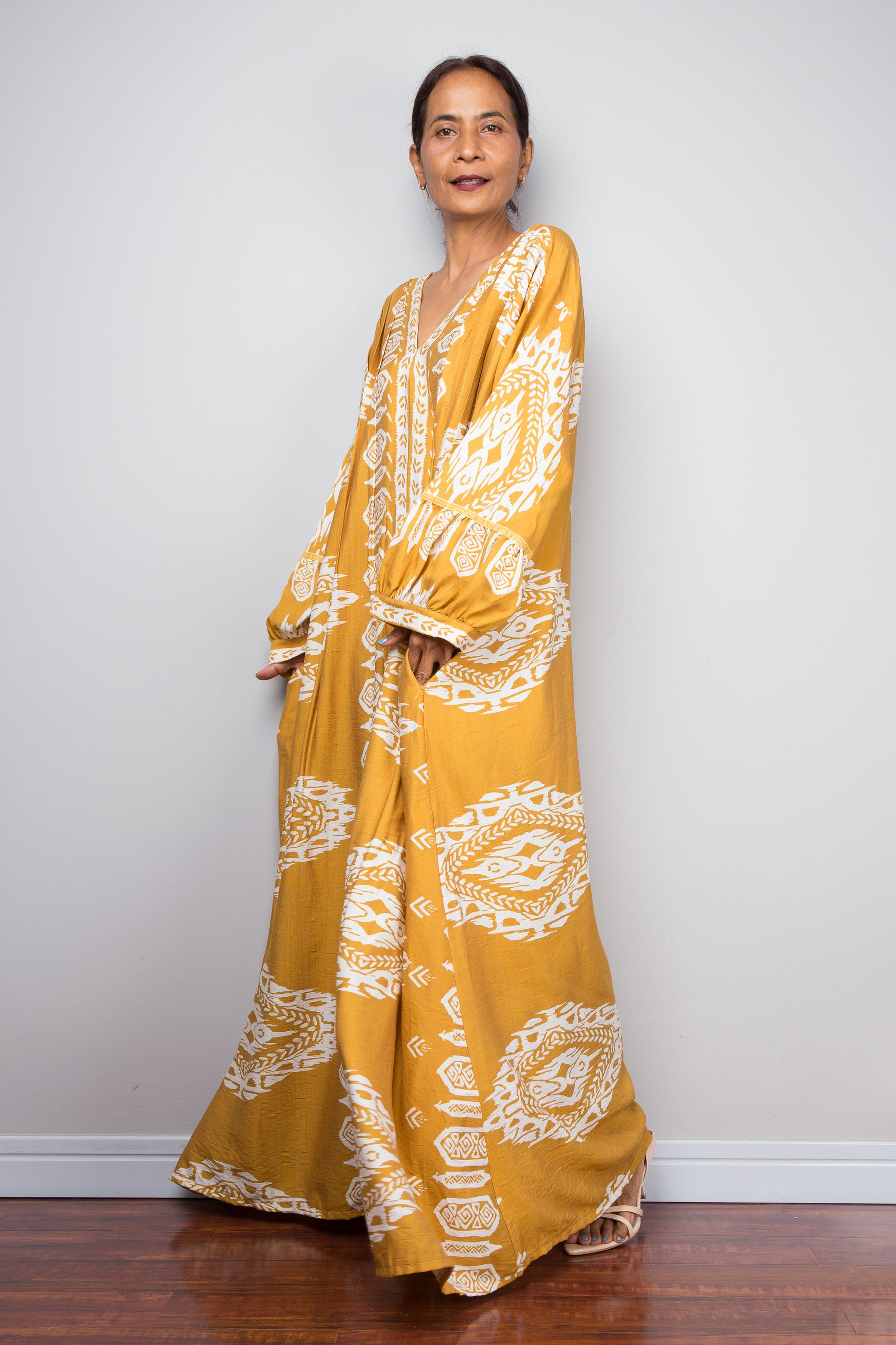 Full-length three-quarter view of a petite model wearing the Nuichan bohemian kaftan maxi dress in mustard yellow and white, showcasing the flowing A-line silhouette, tribal ikat print, and deep V-neckline.