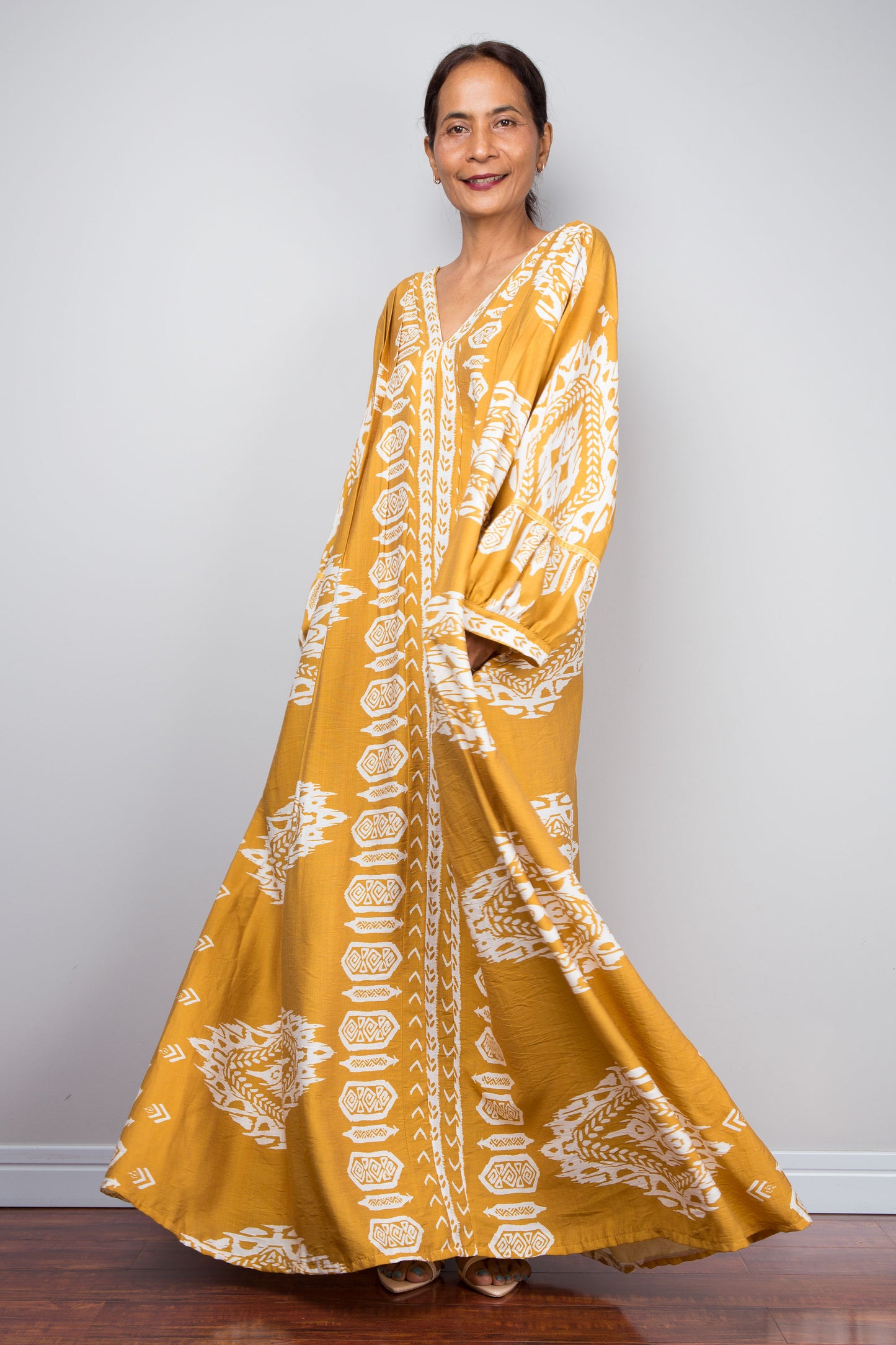 Full-length three-quarter view of a model wearing the Nuichan bohemian kaftan maxi dress in mustard yellow and white, showcasing the flowing A-line silhouette, tribal ikat print, and deep V-neckline.