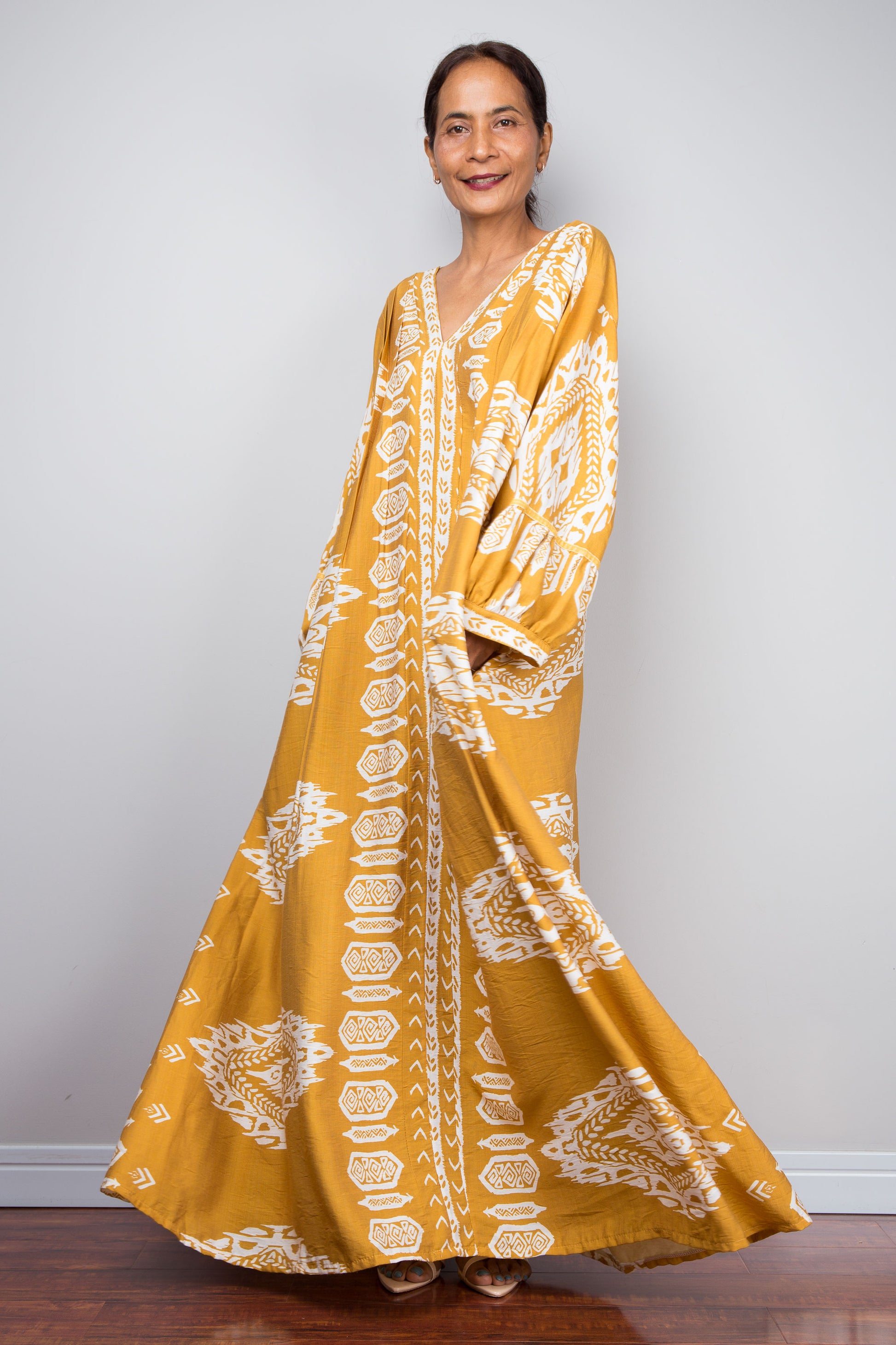 Full-length three-quarter view of a model wearing the Nuichan bohemian kaftan maxi dress in mustard yellow and white, showcasing the flowing A-line silhouette, tribal ikat print, and deep V-neckline.