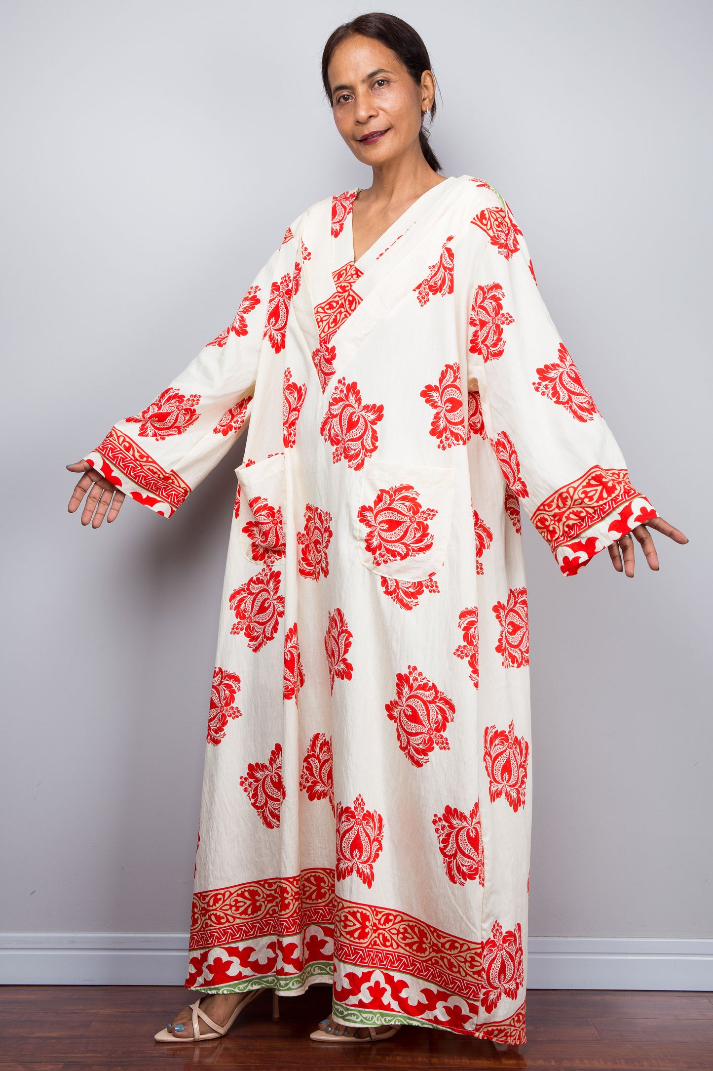 Woman wearing an off white bohemian caftan dress with red floral patterns against a plain background