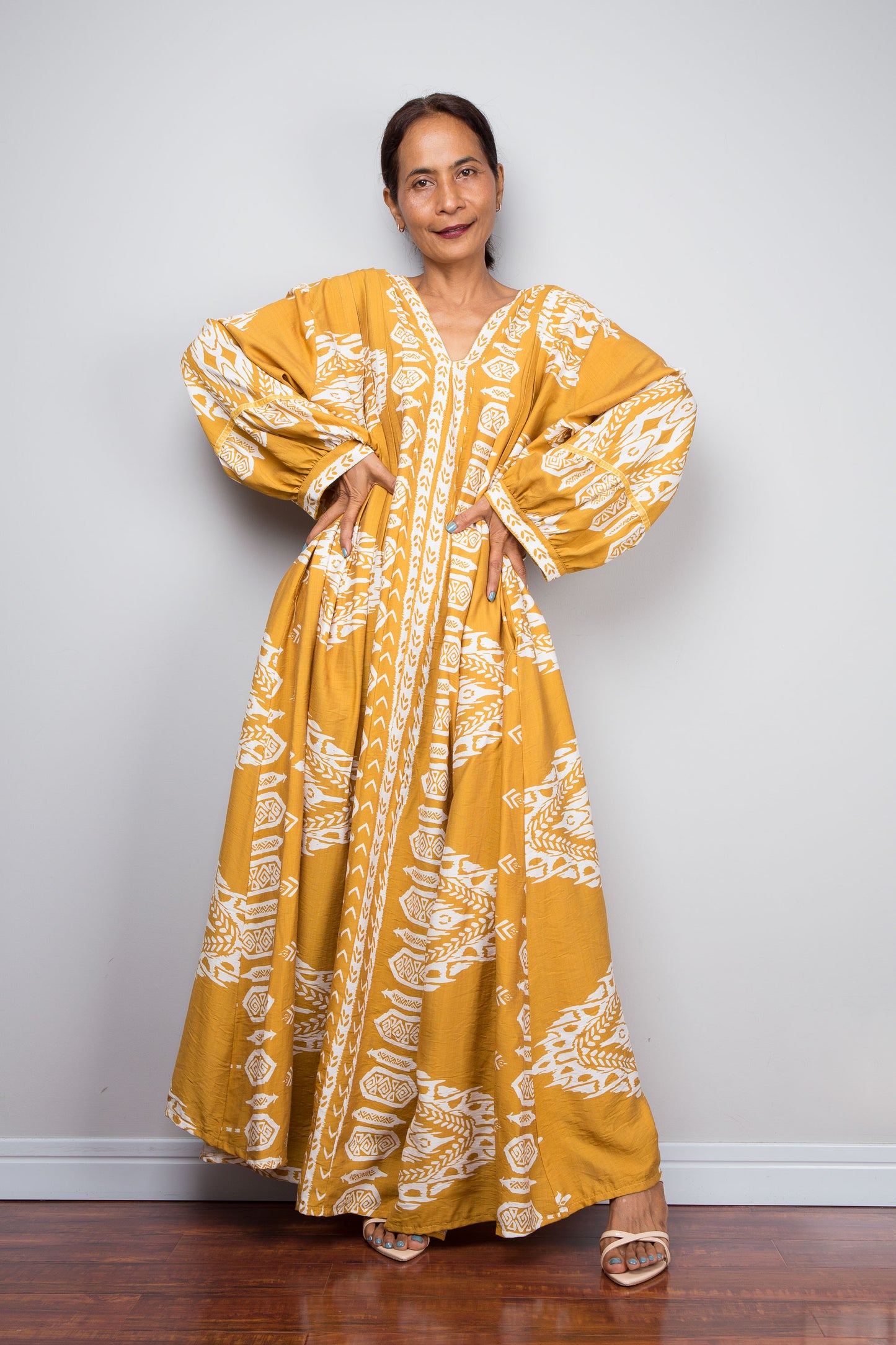 Front-facing studio shot of a model in the Nuichan mustard yellow bohemian caftan dress with hands on hips, highlighting the voluminous bishop sleeves with gathered cuffs and bold white tribal print.