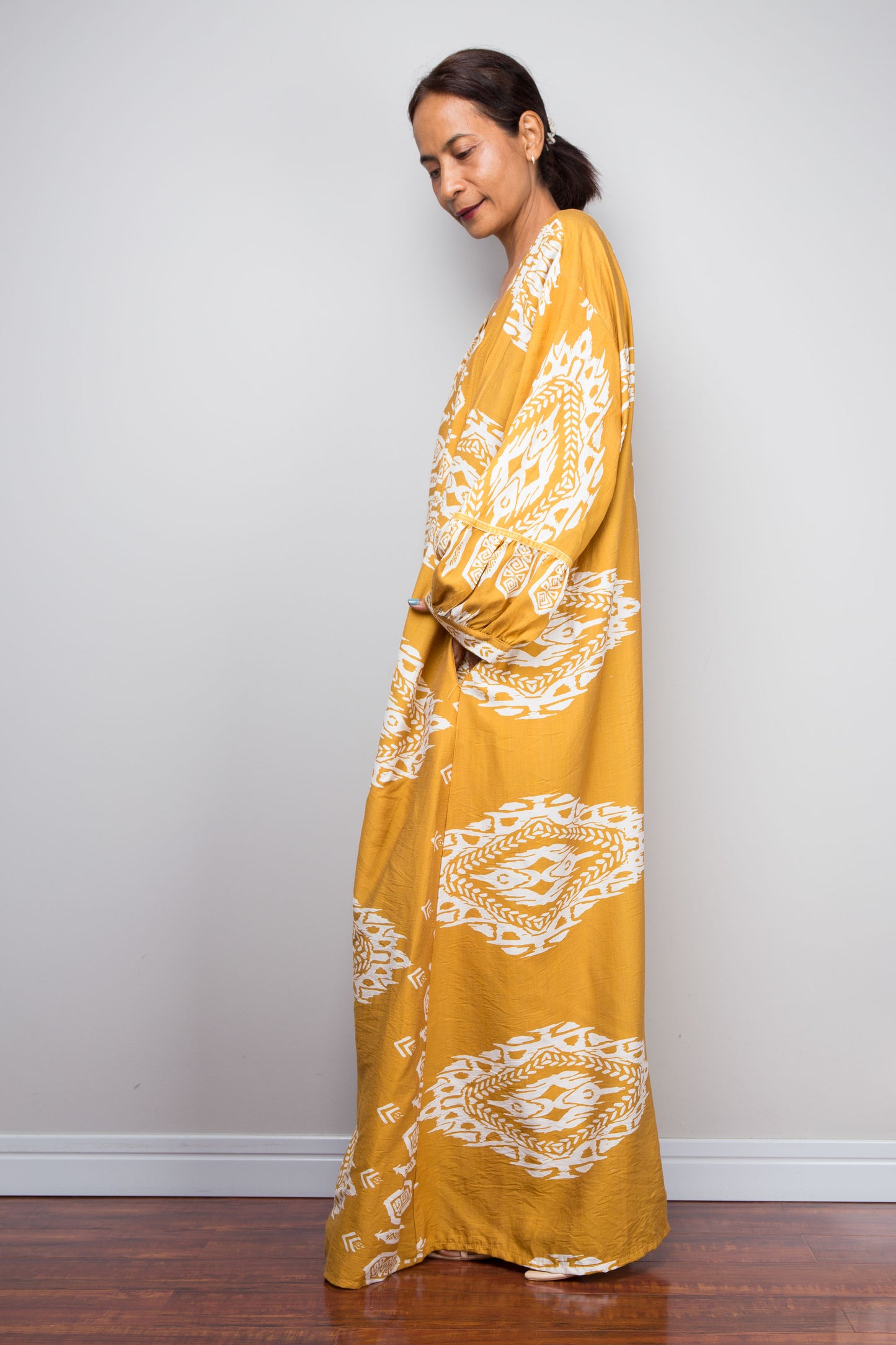 Side profile view of a model wearing the Nuichan mustard and white ikat kaftan dress, emphasising the dramatic floor-length sweep, gathered sleeve cuffs, pockets and intricate placement border print.