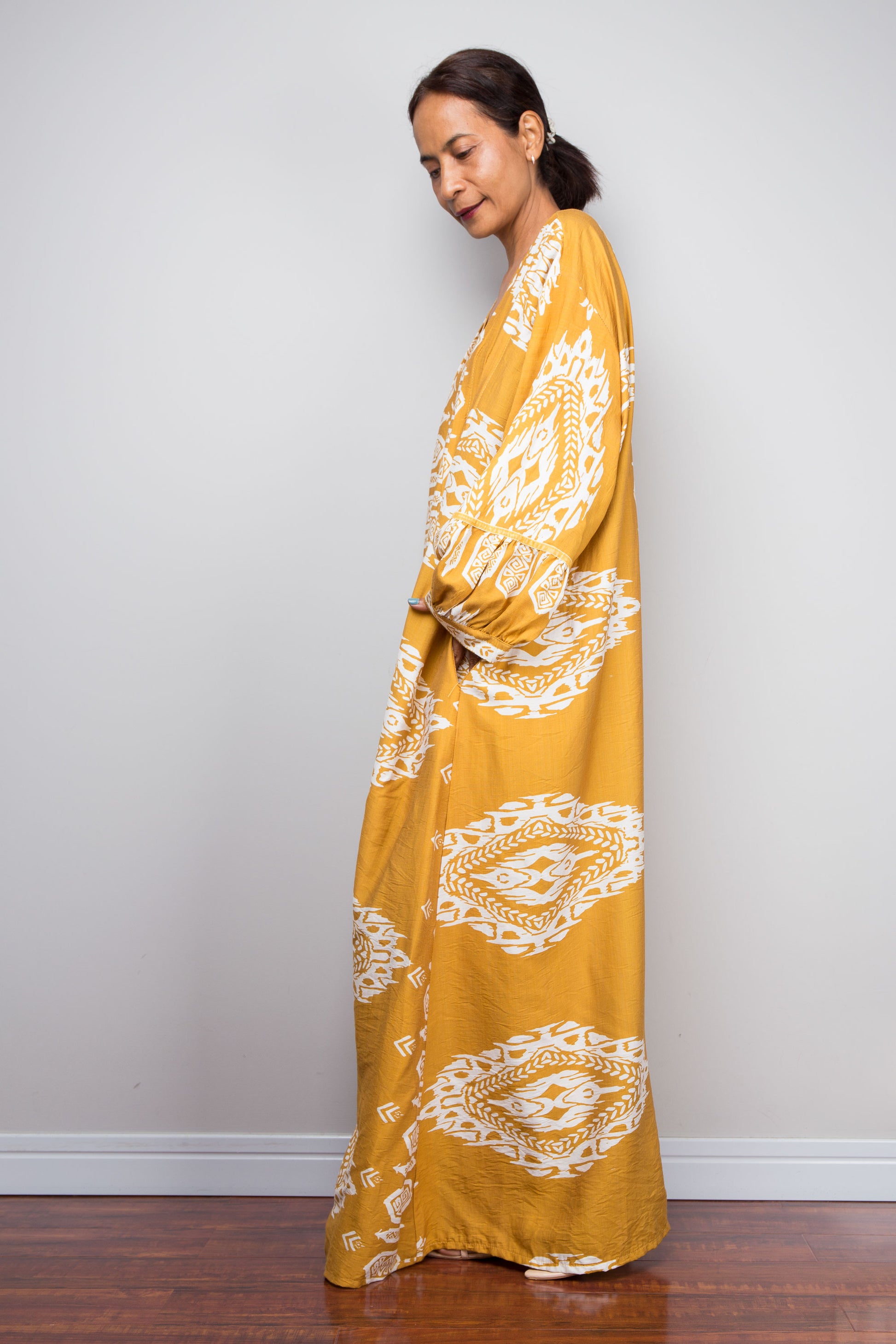 Side profile view of a model wearing the Nuichan mustard and white ikat kaftan dress, emphasising the dramatic floor-length sweep, gathered sleeve cuffs, pockets and intricate placement border print.