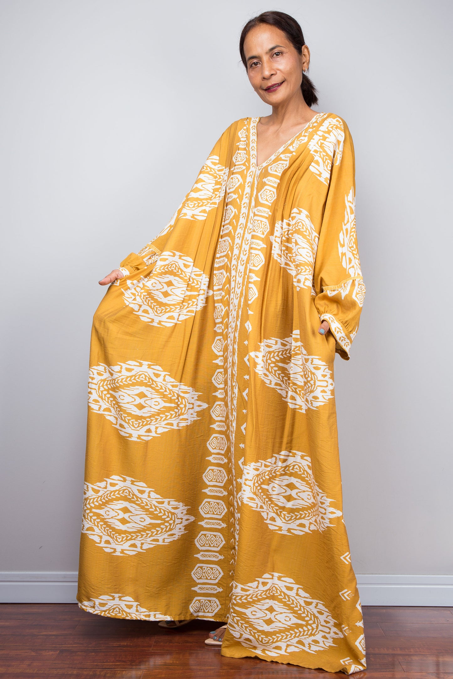 Full-length view of a model wearing the Nuichan bohemian kaftan maxi dress in mustard yellow and white, showcasing the flowing A-line silhouette, tribal ikat print, pockets and deep V-neckline.