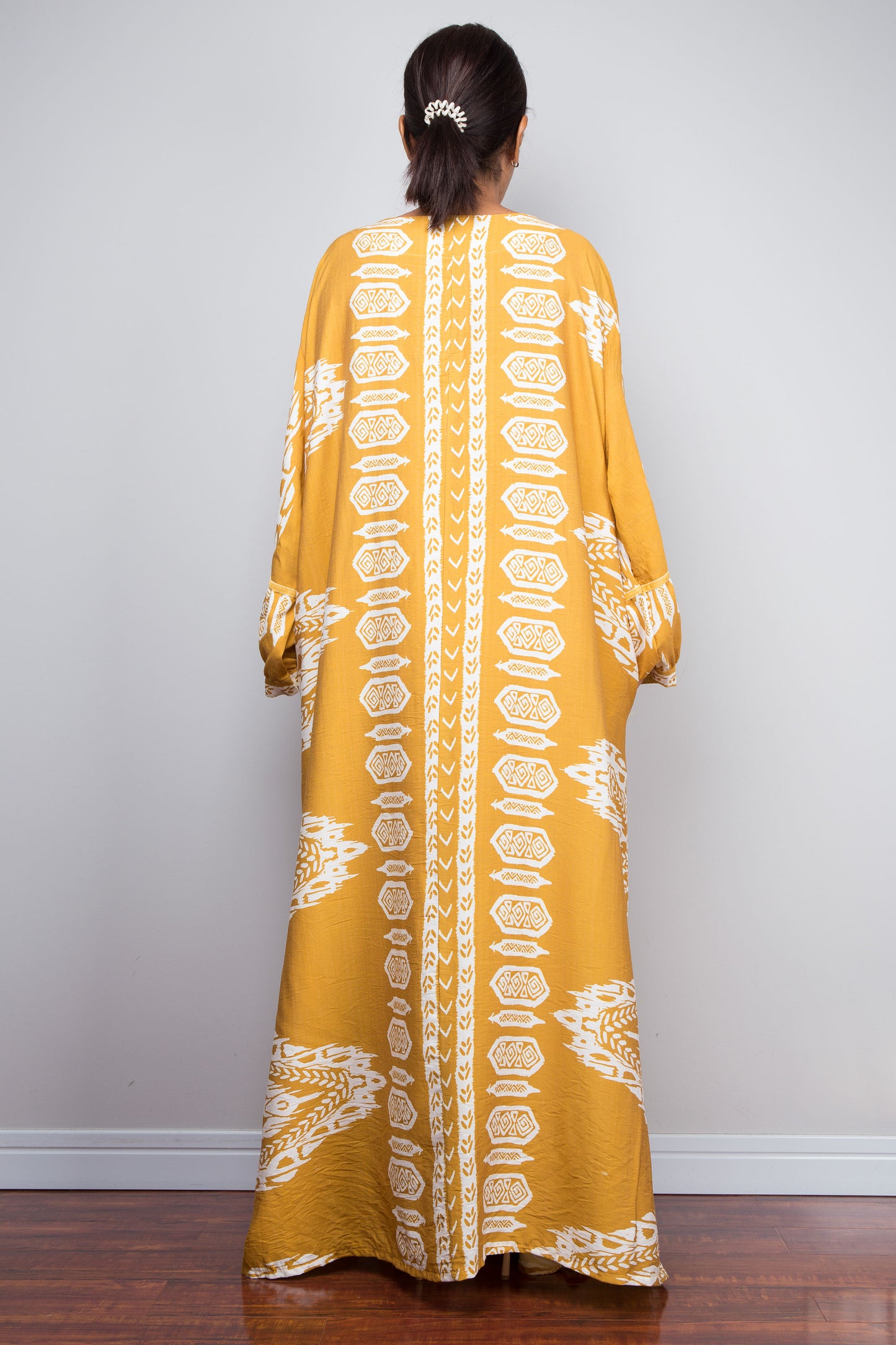 Back profile view of a model wearing the Nuichan mustard and white ikat kaftan dress, emphasising the dramatic floor-length sweep, gathered sleeve cuffs, and intricate placement border print.