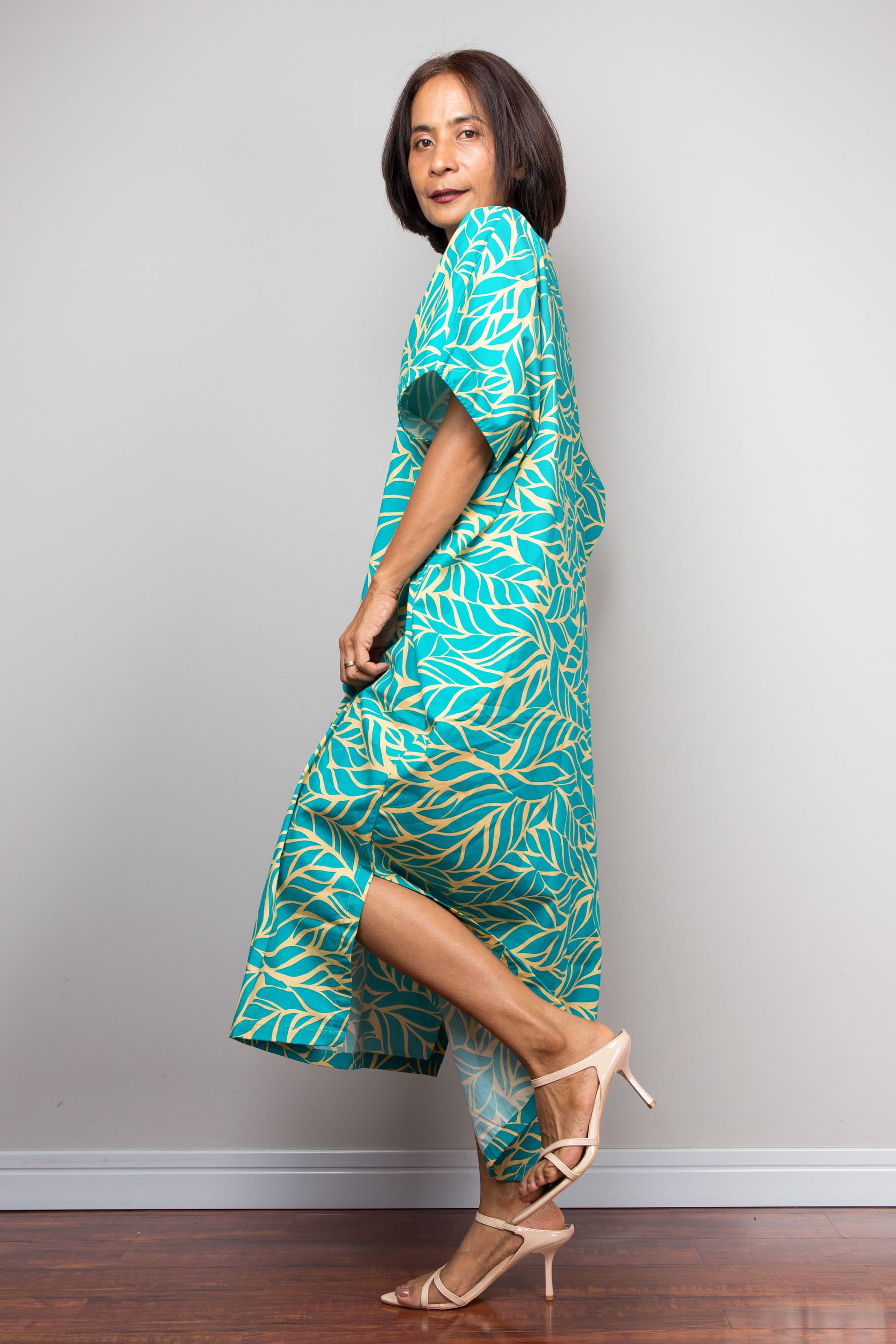 Petite Woman wearing an aqua green and yellow patterned kaftan dress against a gray wall.
