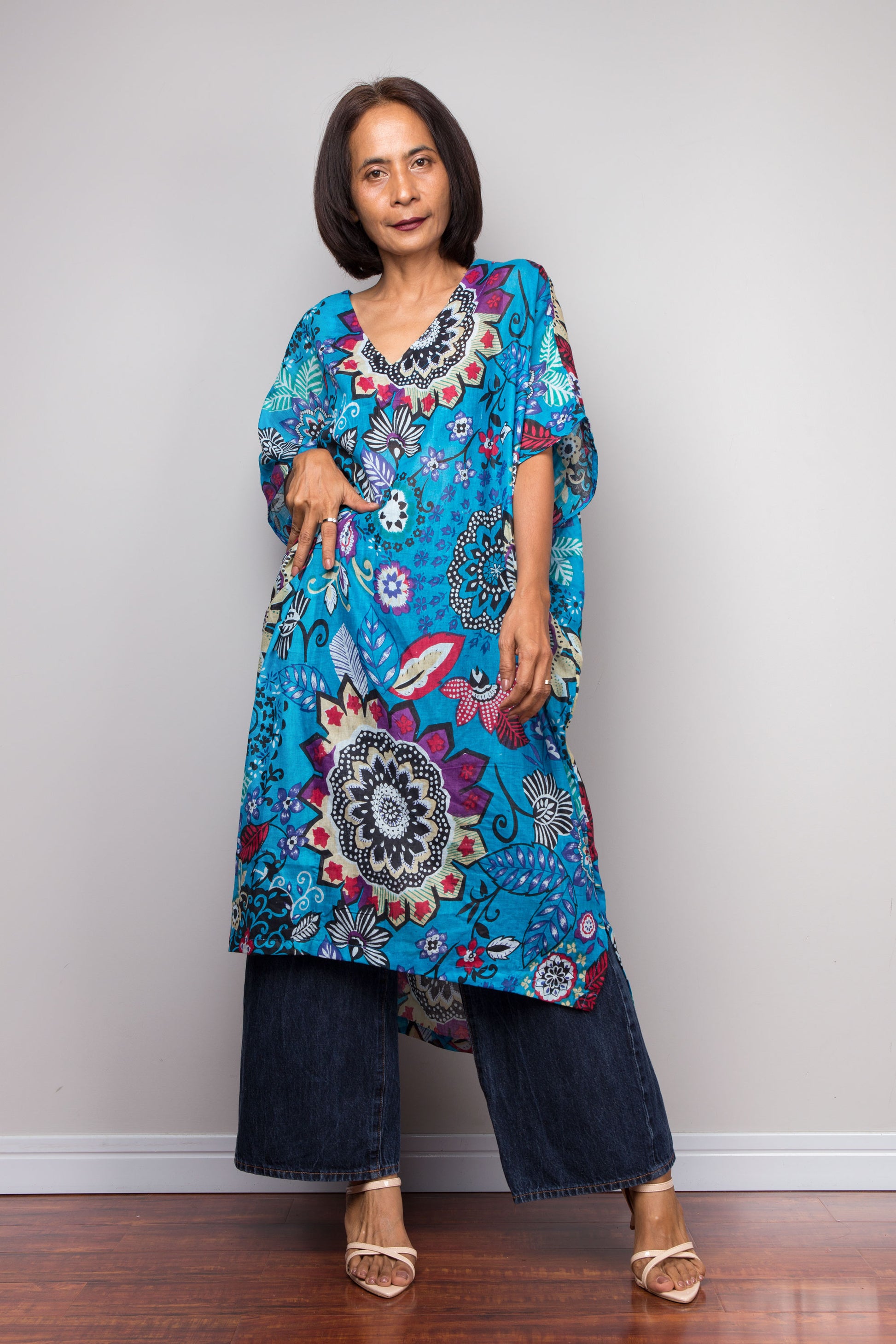 Woman wearing a colorful floral kaftan dress with blue pants against a plain background