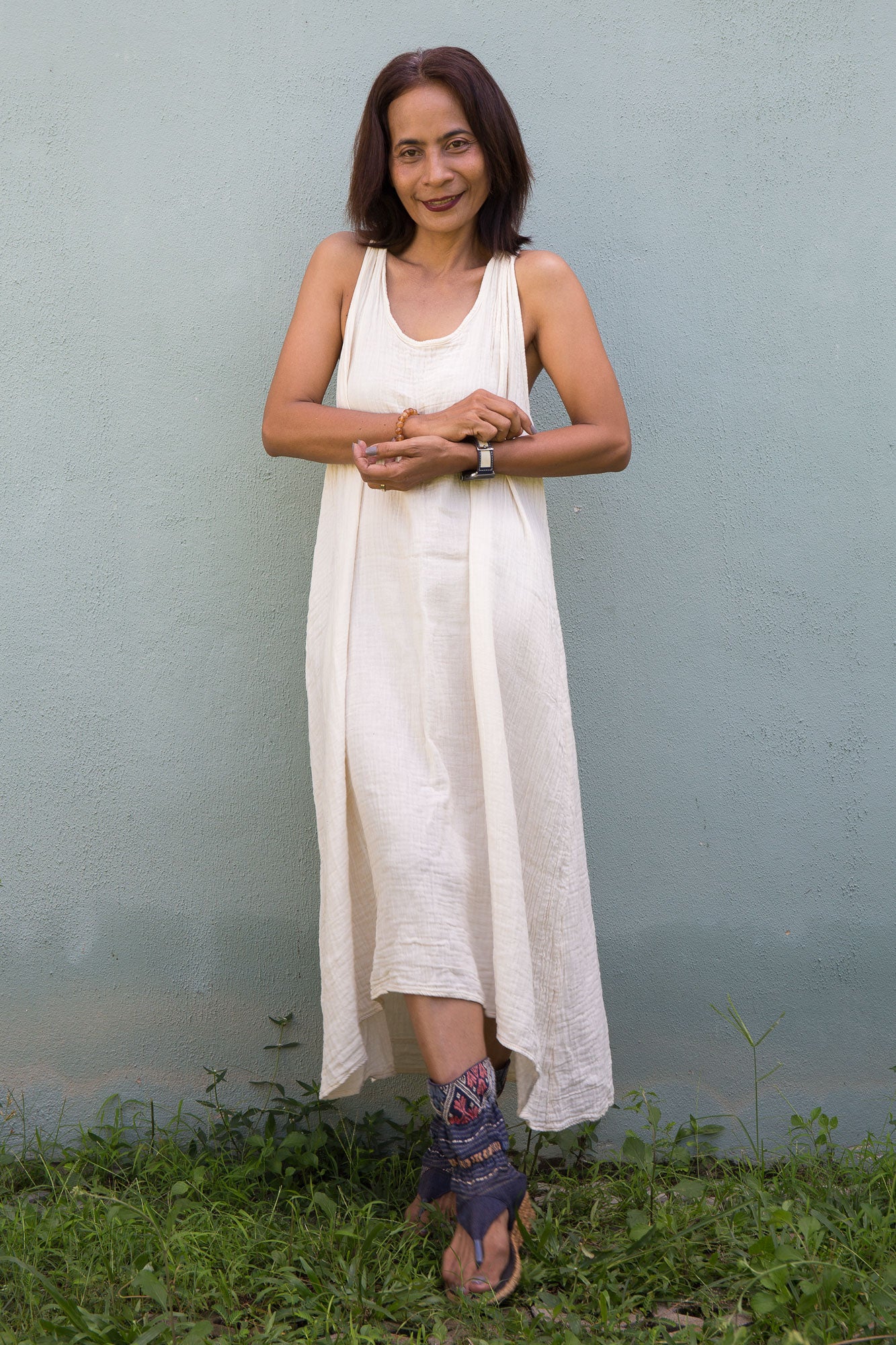 Woman in a white cotton gauze dress standing in front of a gray wall with green grass at the bottom.