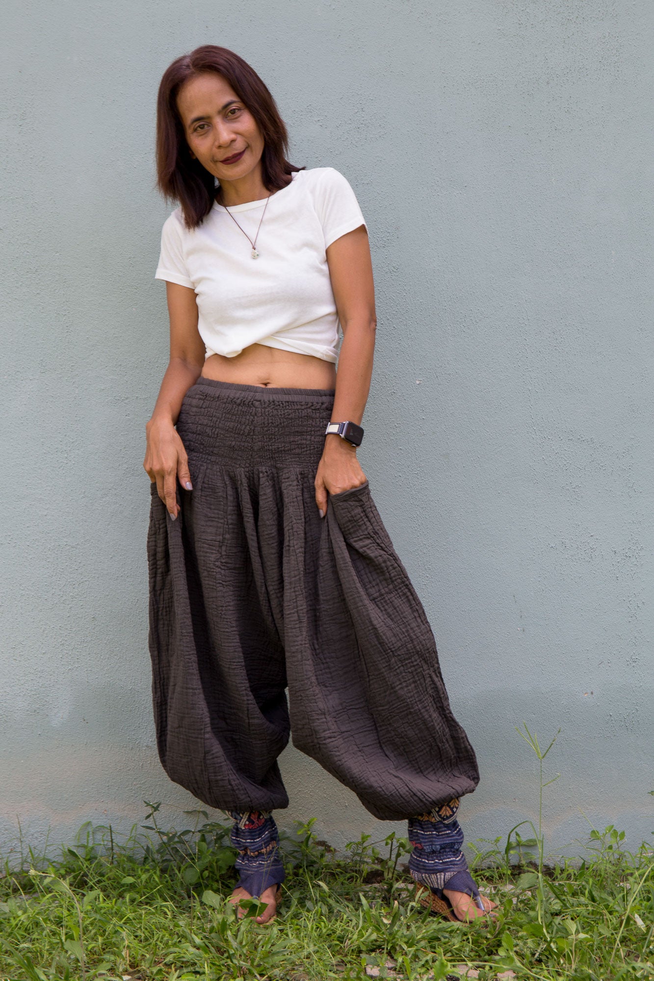 Woman wearing a white top and gray wide-leg cotton harem pants standing against a gray wall.