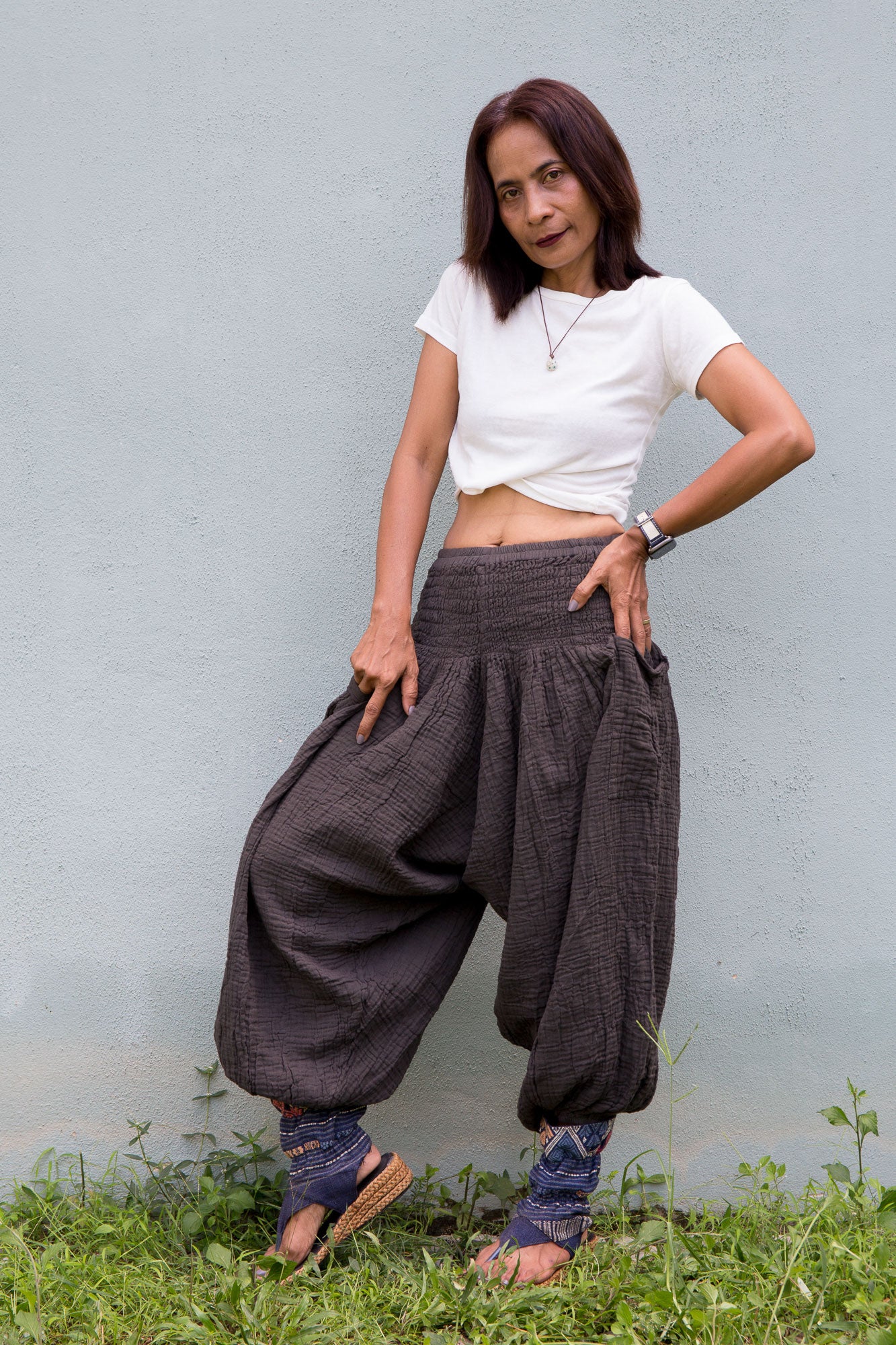 Woman wearing a white top and charcoal grey cotton harem pants standing against a gray wall.
