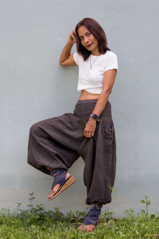 Woman in white top and grey wide-leg harem pants standing against a light gray wall with grass at the bottom.