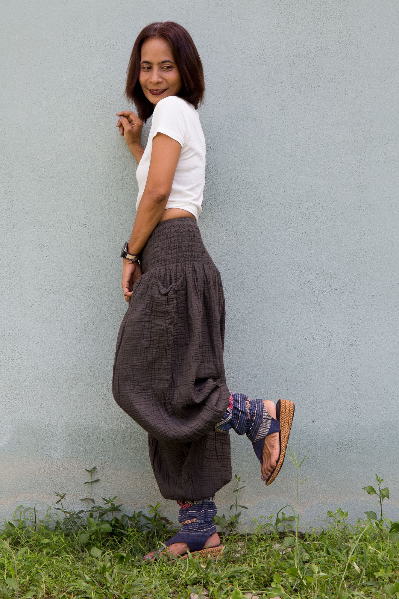 Woman wearing a white top and cotton gauze baggy harem pants standing against a light gray wall.