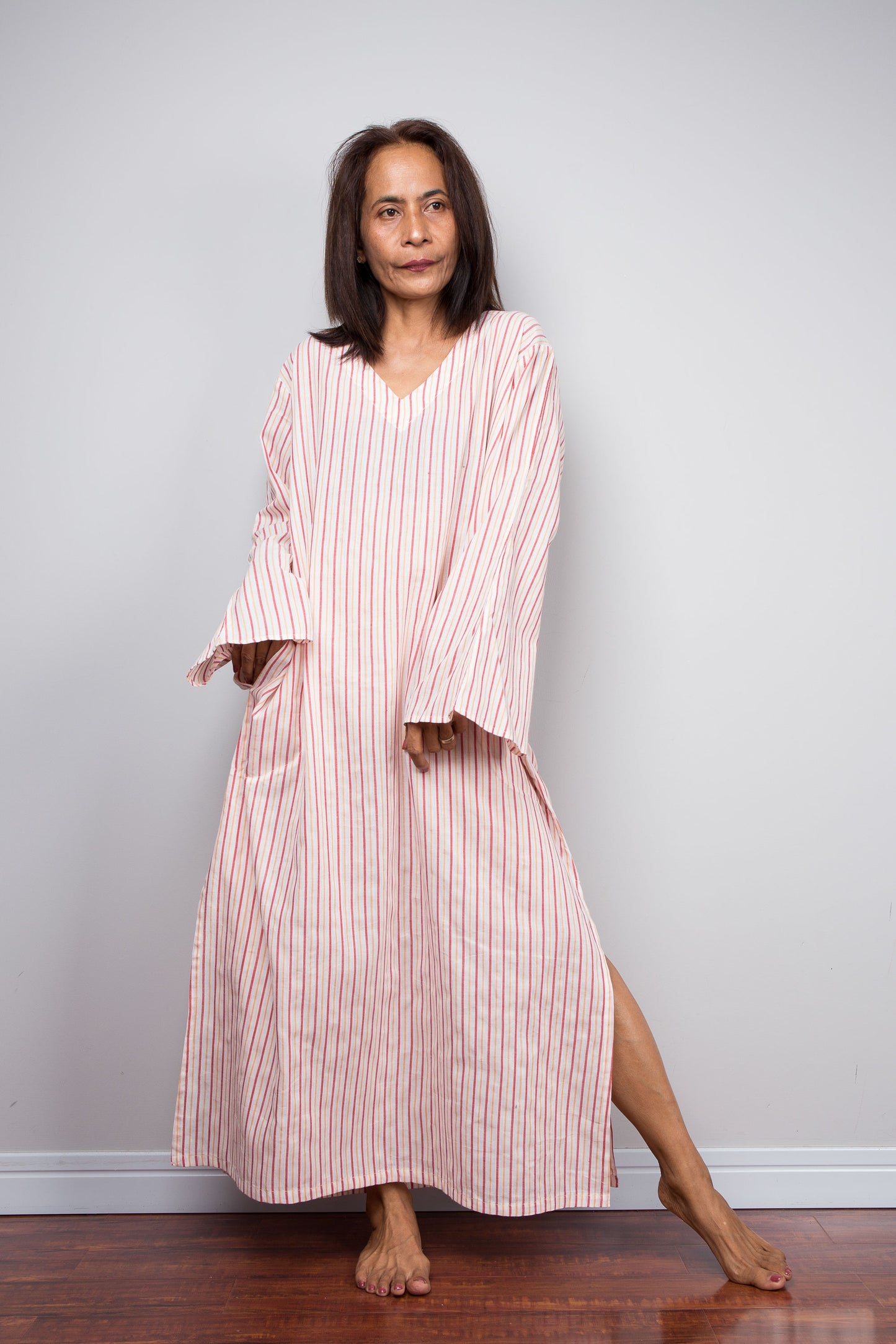 A petite woman wearing a pink and white vertically striped kaftan dress with wide bell sleeves, standing barefoot on a wooden floor against a light gray wall.