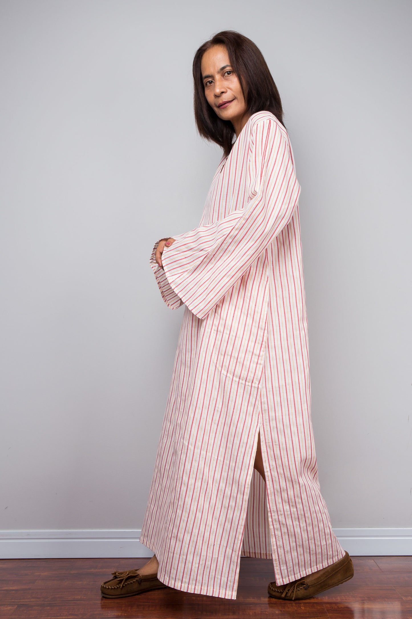A woman modeling a long-sleeved, floor-length pink and white striped robe or abaya with brown slip-on shoes, standing against a light gray wall and dark wood floor.