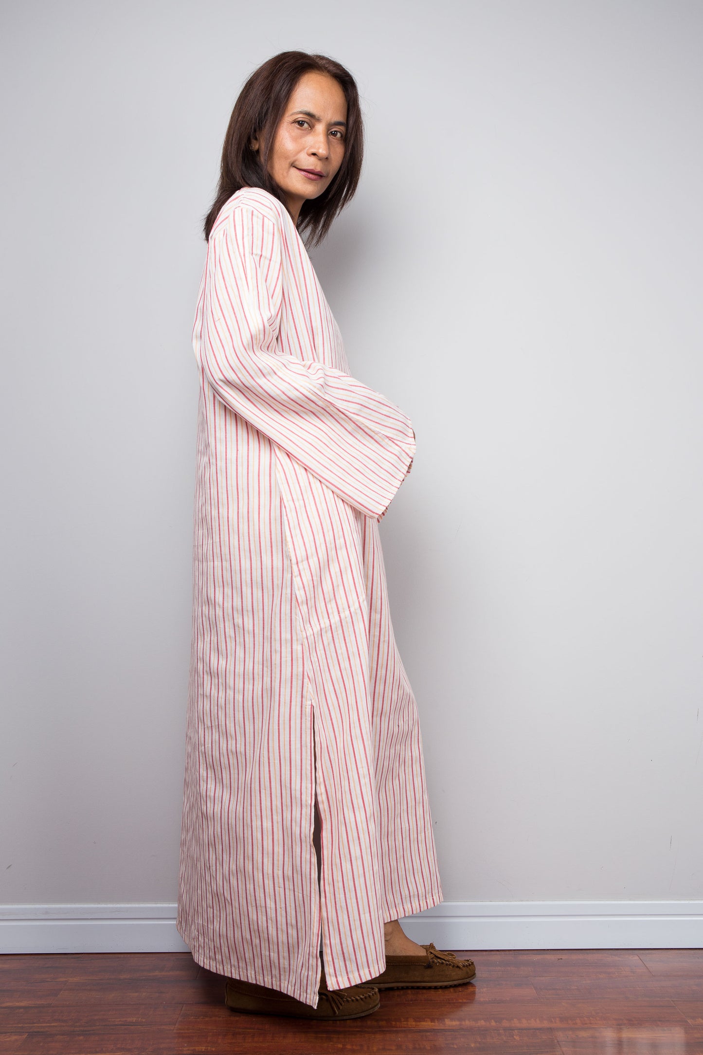 A woman wearing a long-sleeved, floor-length pink and white striped robe or kimono-style garment, standing in profile against a plain light gray wall on a wooden floor.
