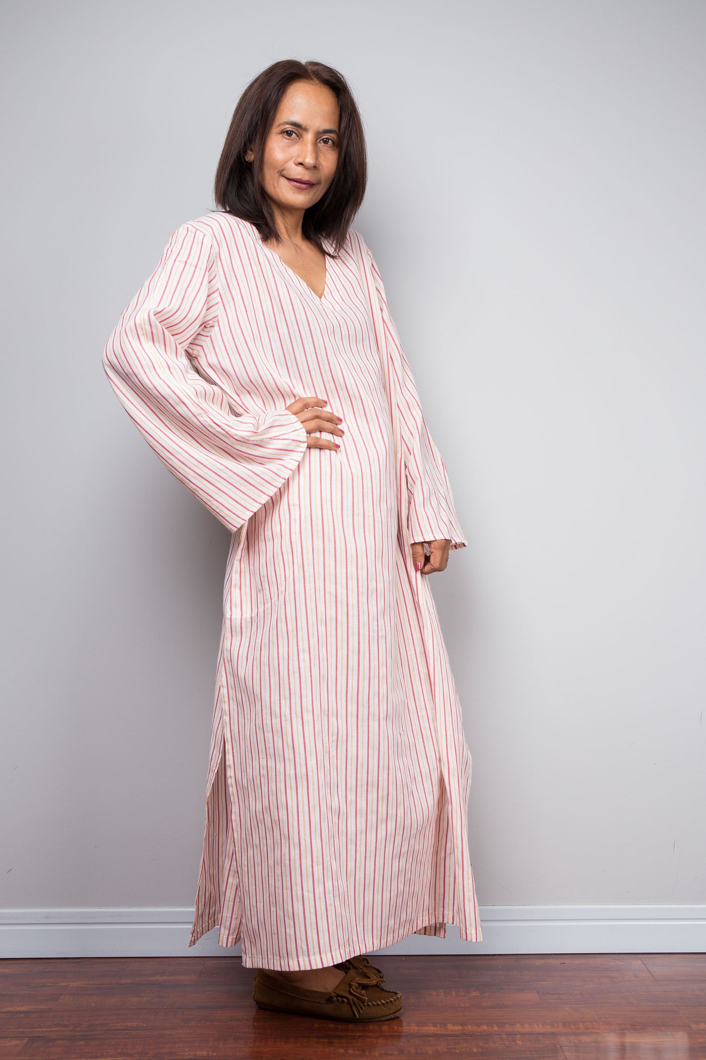 A woman modeling a long-sleeved, V-neck maxi dress with vertical pink and white stripes, standing against a light gray wall on a dark wood floor.