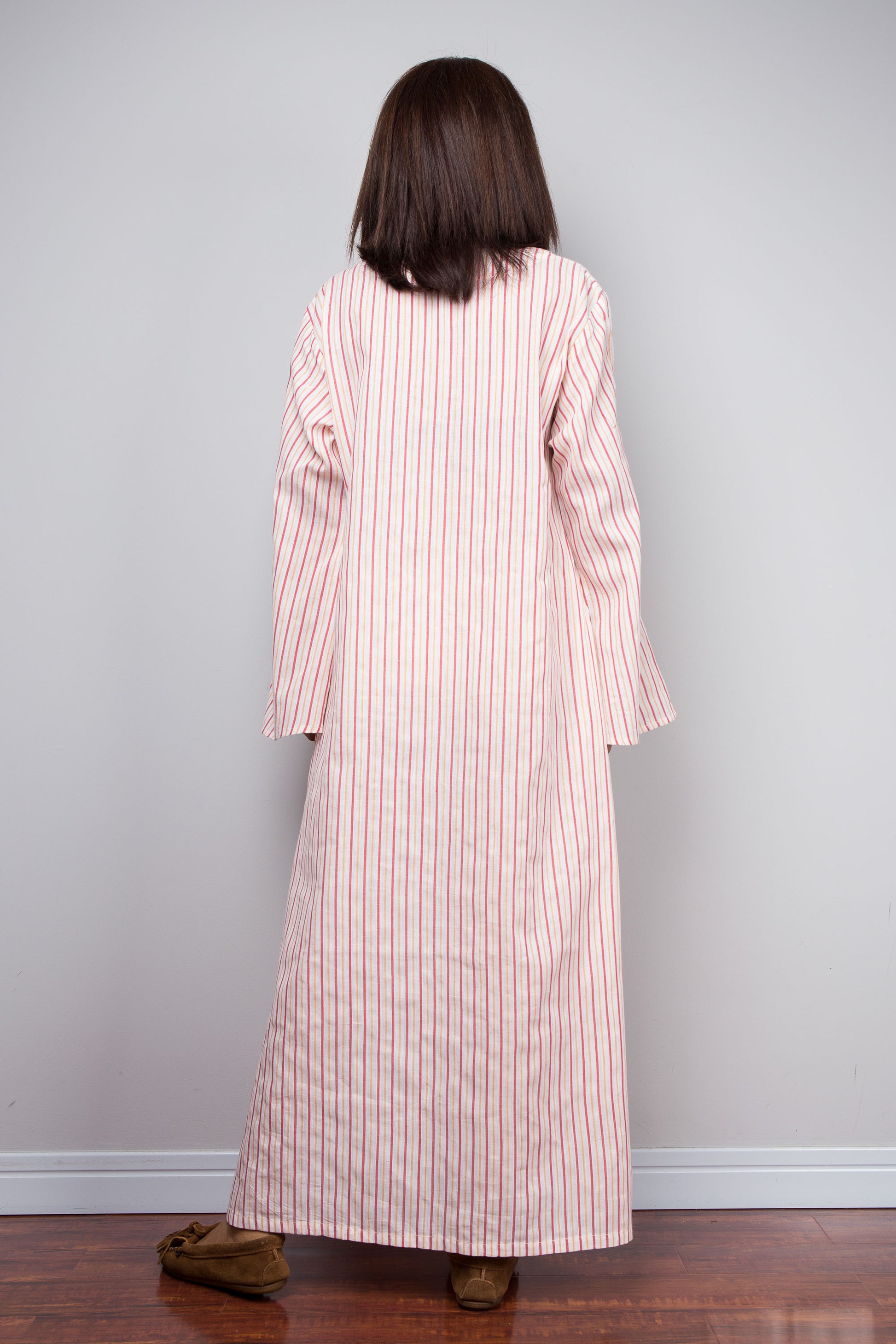 A person viewed from behind wearing a long-sleeved, floor-length striped robe or nightgown in pink and white, standing on a wooden floor against a plain light gray wall.