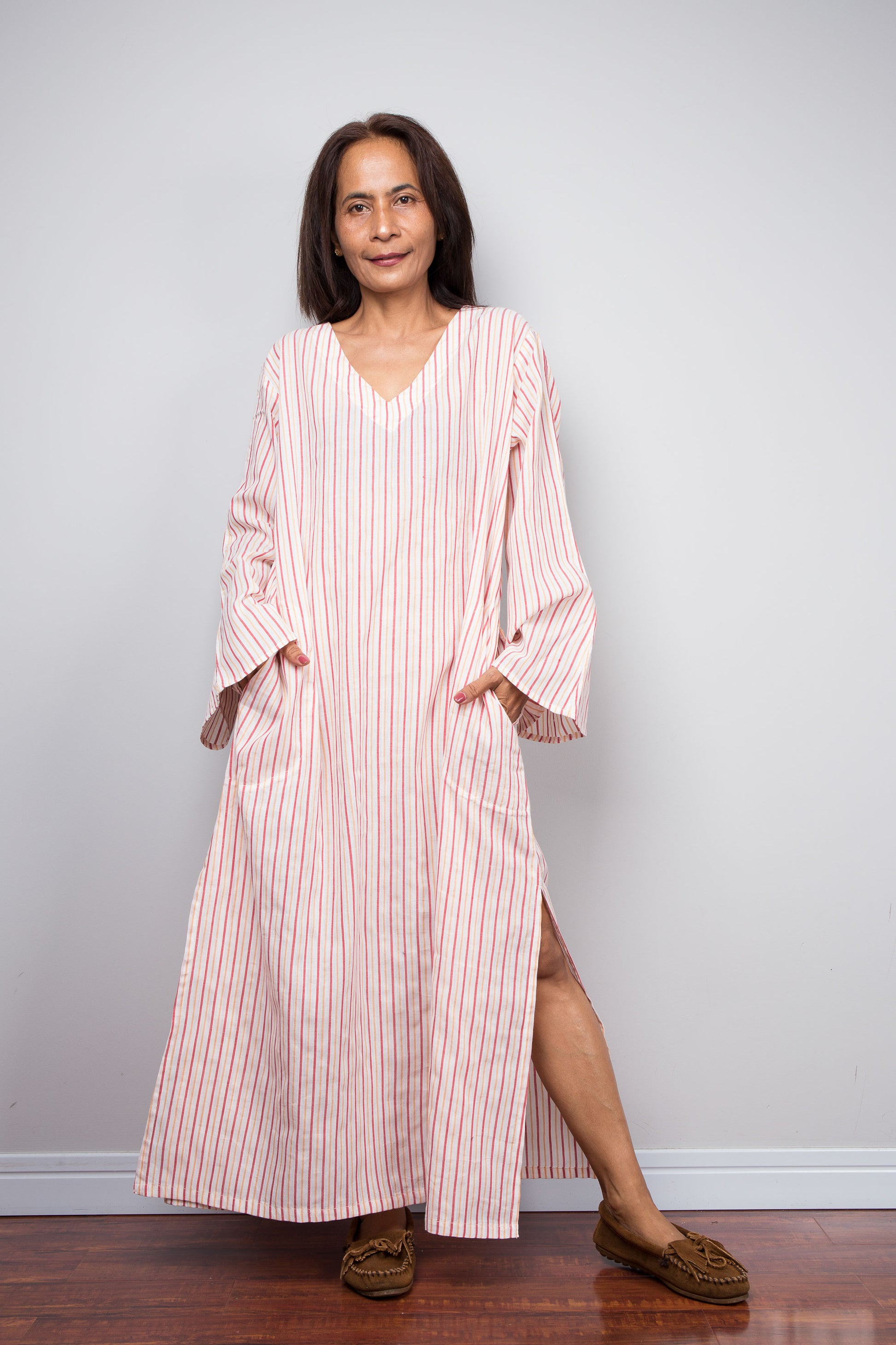 A woman wearing a long-sleeved, V-neck petite kaftan dress with vertical pink and white stripes, standing against a light gray wall on a wooden floor.