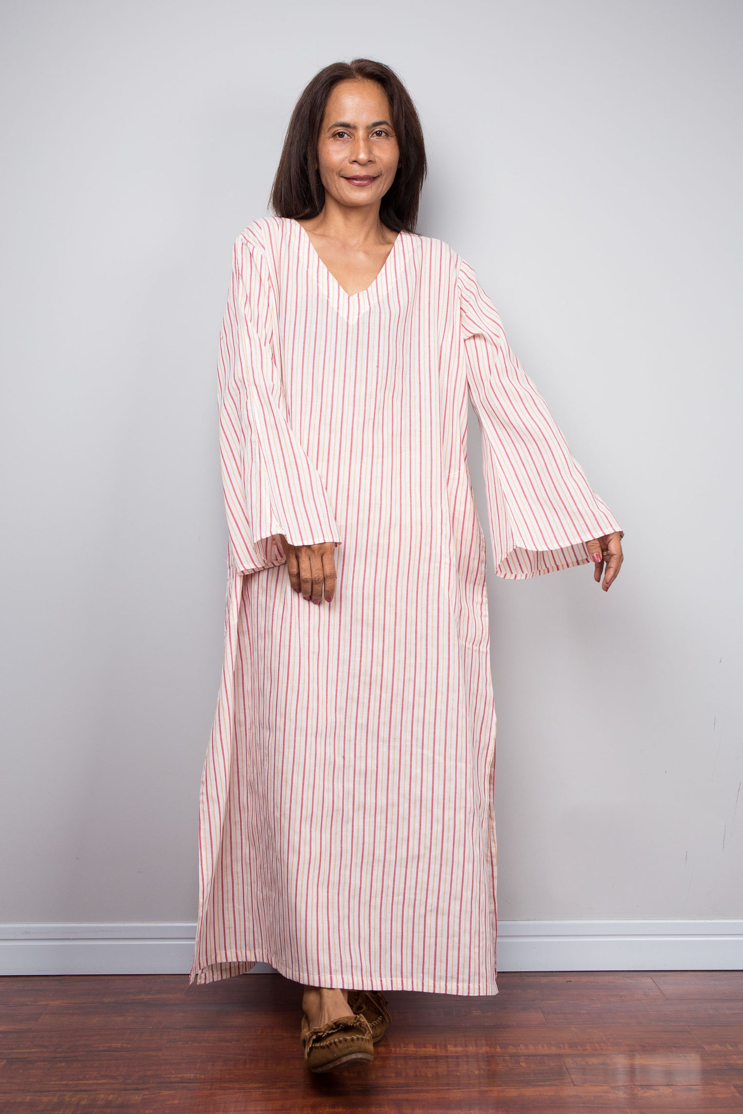 A petite woman modeling a long-sleeved, V-neck kaftan dress with vertical pink and white stripes, standing against a light gray wall and dark wood floor.