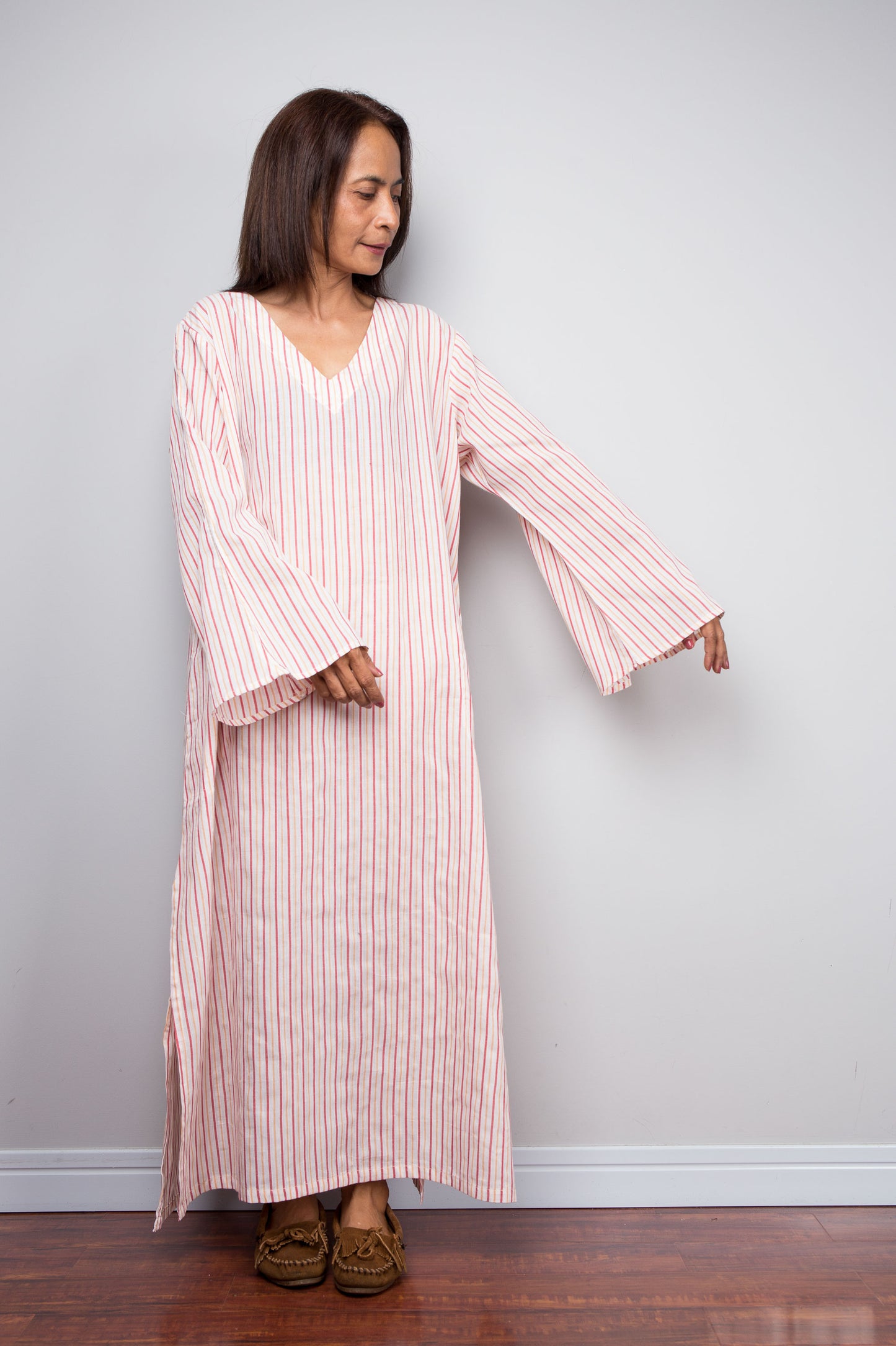 A woman modeling a long-sleeved, V-neck petite kaftan dress with vertical pink and white stripes, standing against a plain light gray wall on a wooden floor.