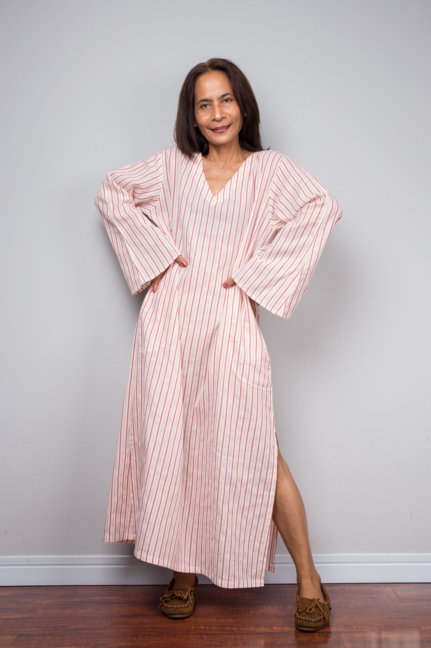 A petite woman modeling a long-sleeved, V-neck maxi kaftan, for petite, with pink and white vertical stripes, standing against a light gray wall on a wooden floor.