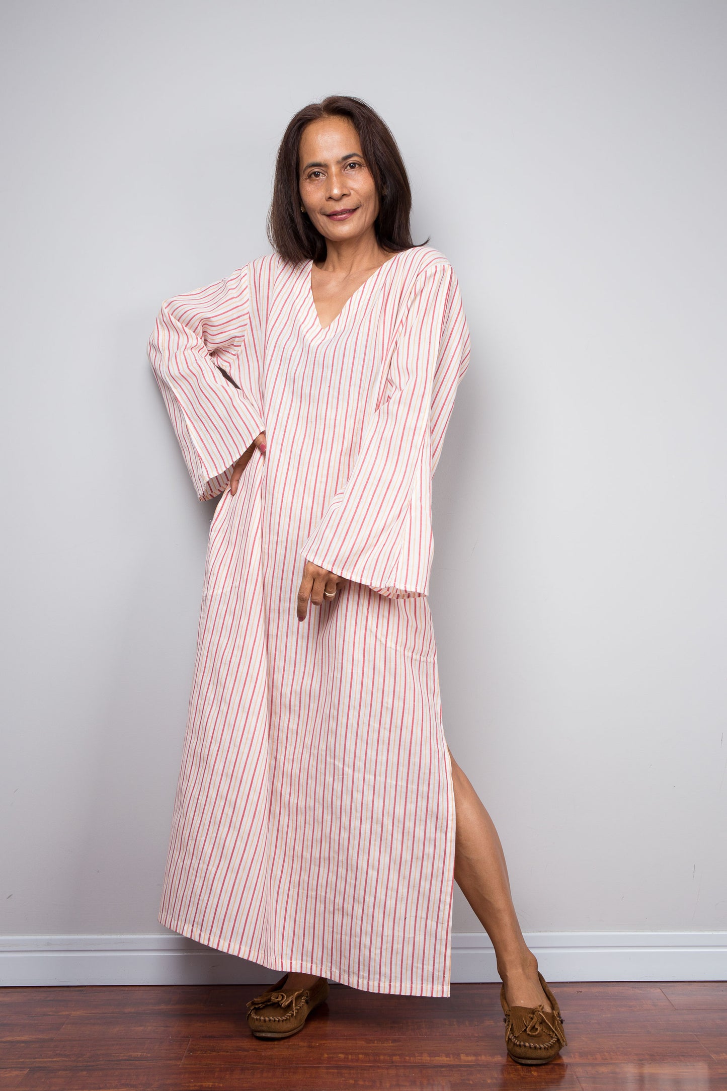 A woman modeling a long bell sleeved, V-neck petite kaftan with thin vertical pink and white stripes, standing against a light gray wall on a dark wood floor.