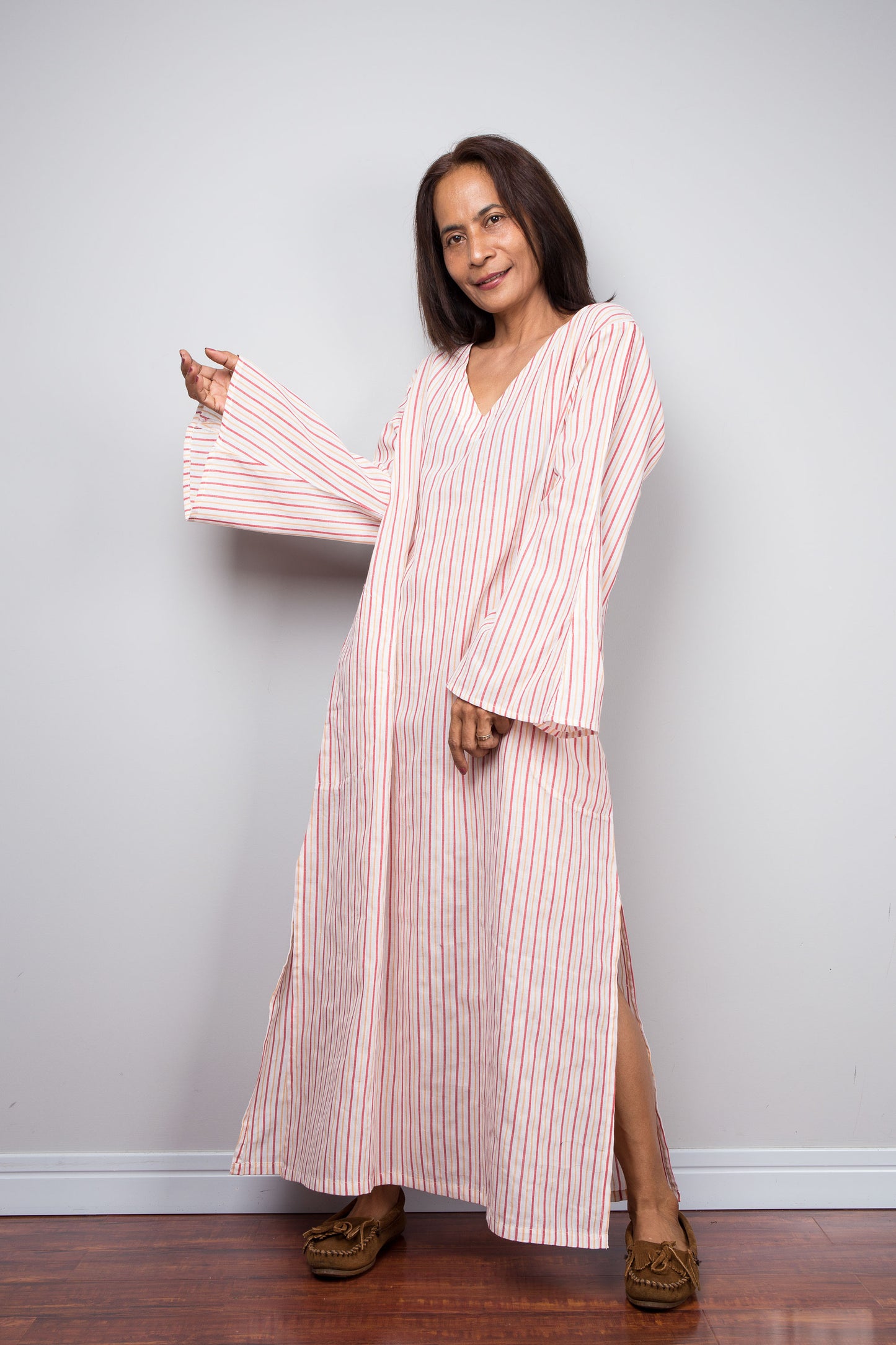 A petite woman modeling a long-sleeved, V-neck cotton kaftan dress with pink and white vertical stripes, standing against a light gray wall on a wooden floor.