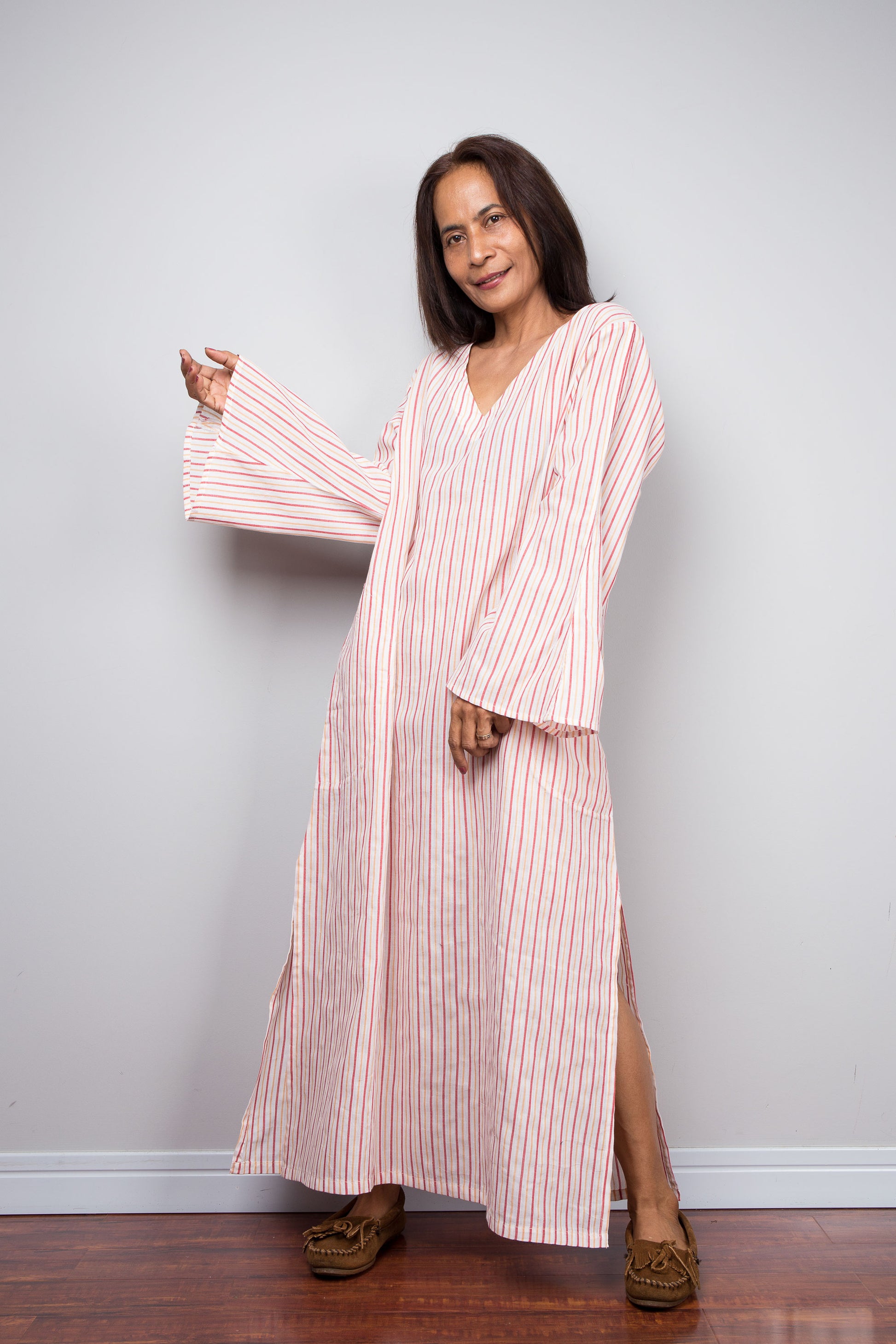 A petite woman modeling a long-sleeved, V-neck cotton kaftan dress with pink and white vertical stripes, standing against a light gray wall on a wooden floor.