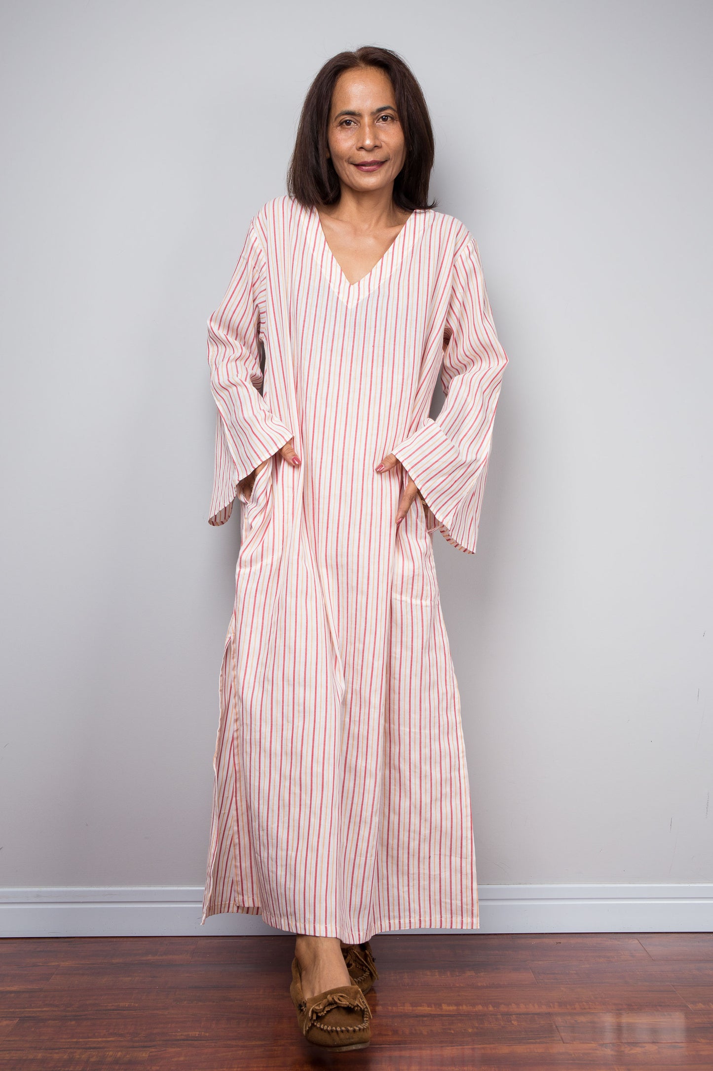 A woman modeling a long-sleeved, V-neck petite caftan dress with pink and white vertical stripes, standing against a light gray wall on a dark wood floor.