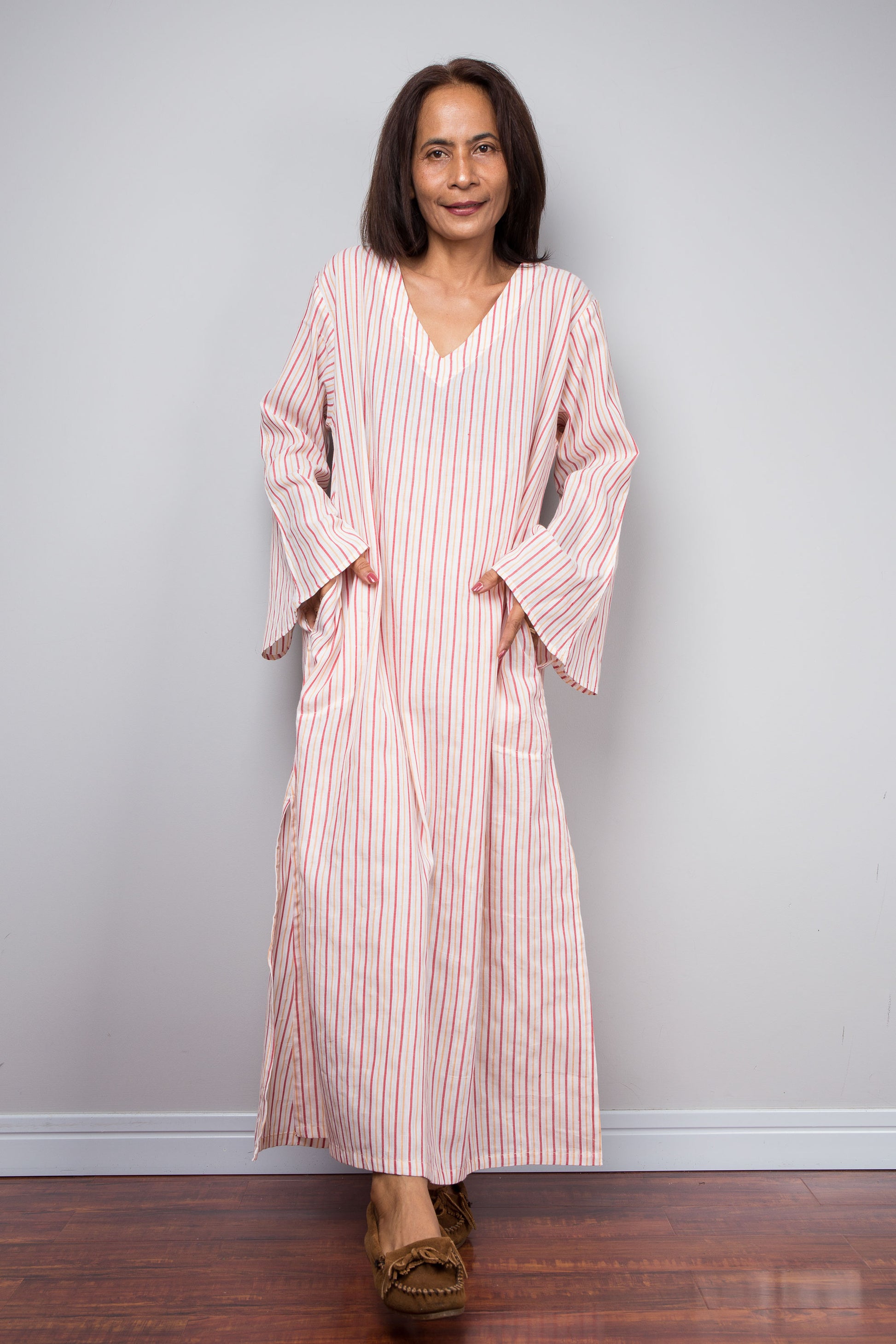 A woman modeling a long-sleeved, V-neck petite caftan dress with pink and white vertical stripes, standing against a light gray wall on a dark wood floor.