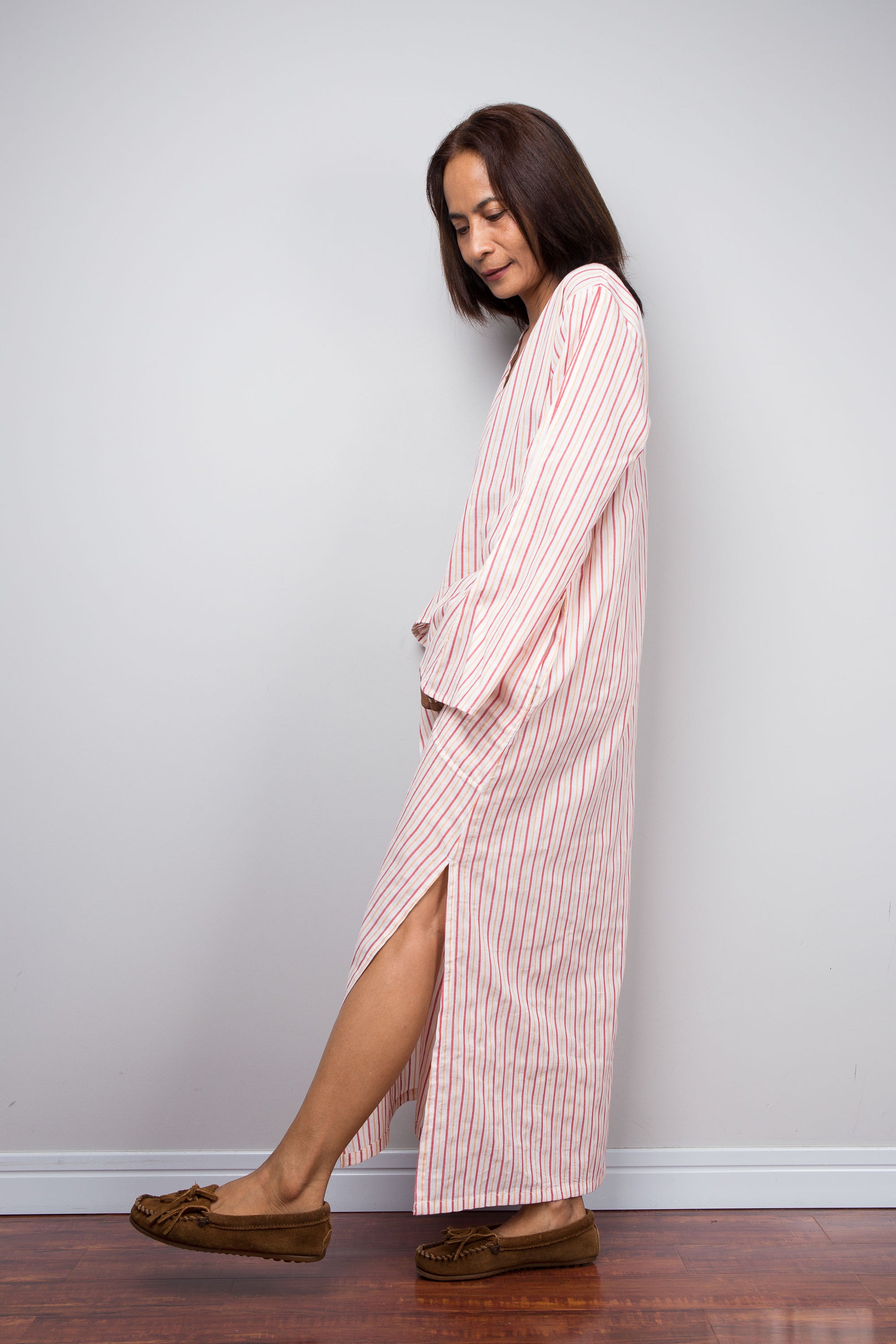 A woman wearing a pink and white vertically striped long robe or nightgown with brown suede moccasin-style flats, standing in profile against a plain light gray wall on a wooden floor.